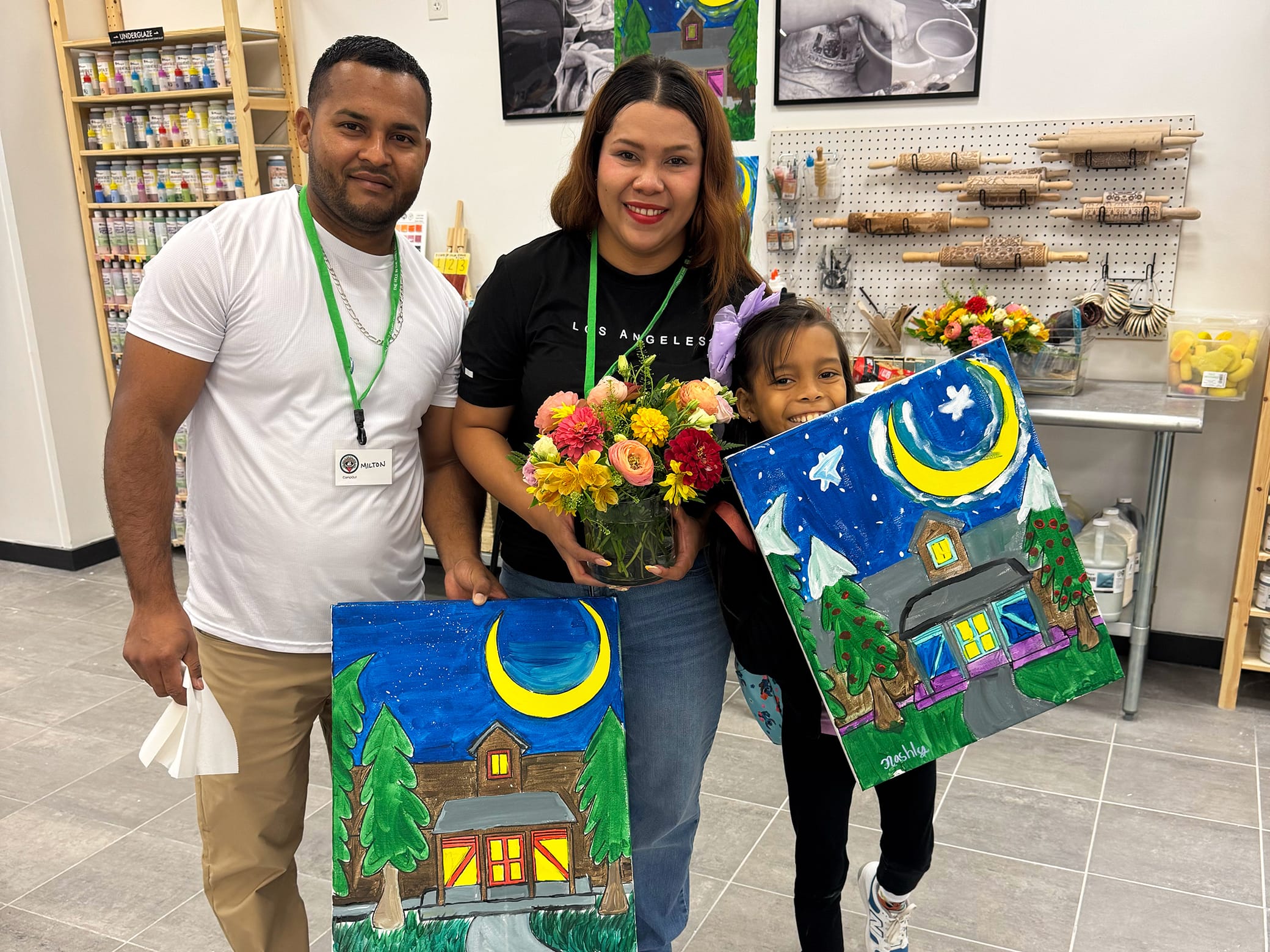 Three people smile indoors, holding paintings of a house with trees and a crescent moon at night. The woman in the middle also holds a bouquet of flowers. Art supplies and paintings are visible in the background.