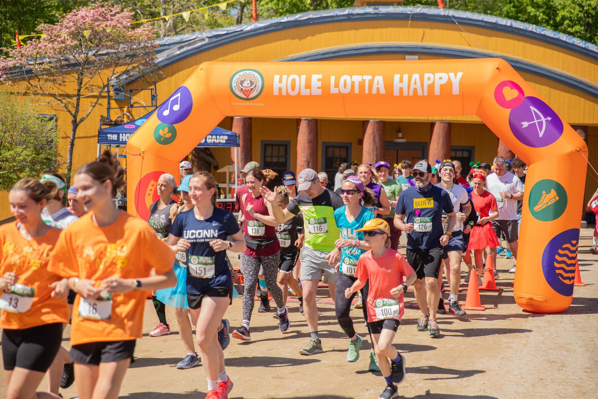 A group of runners starts a race under an orange inflatable arch that reads HOLE LOTTA HAPPY. Some people wear bibs and orange shirts; others are in athletic gear. Trees and a building are in the background.