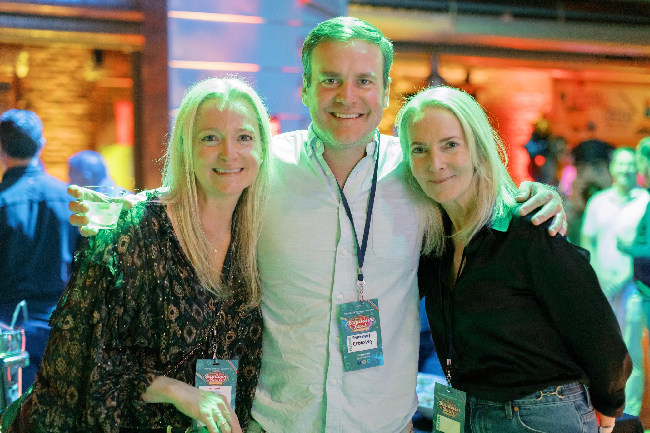 Three people with event badges smile and pose closely together at an indoor event with colorful lighting and a lively background.