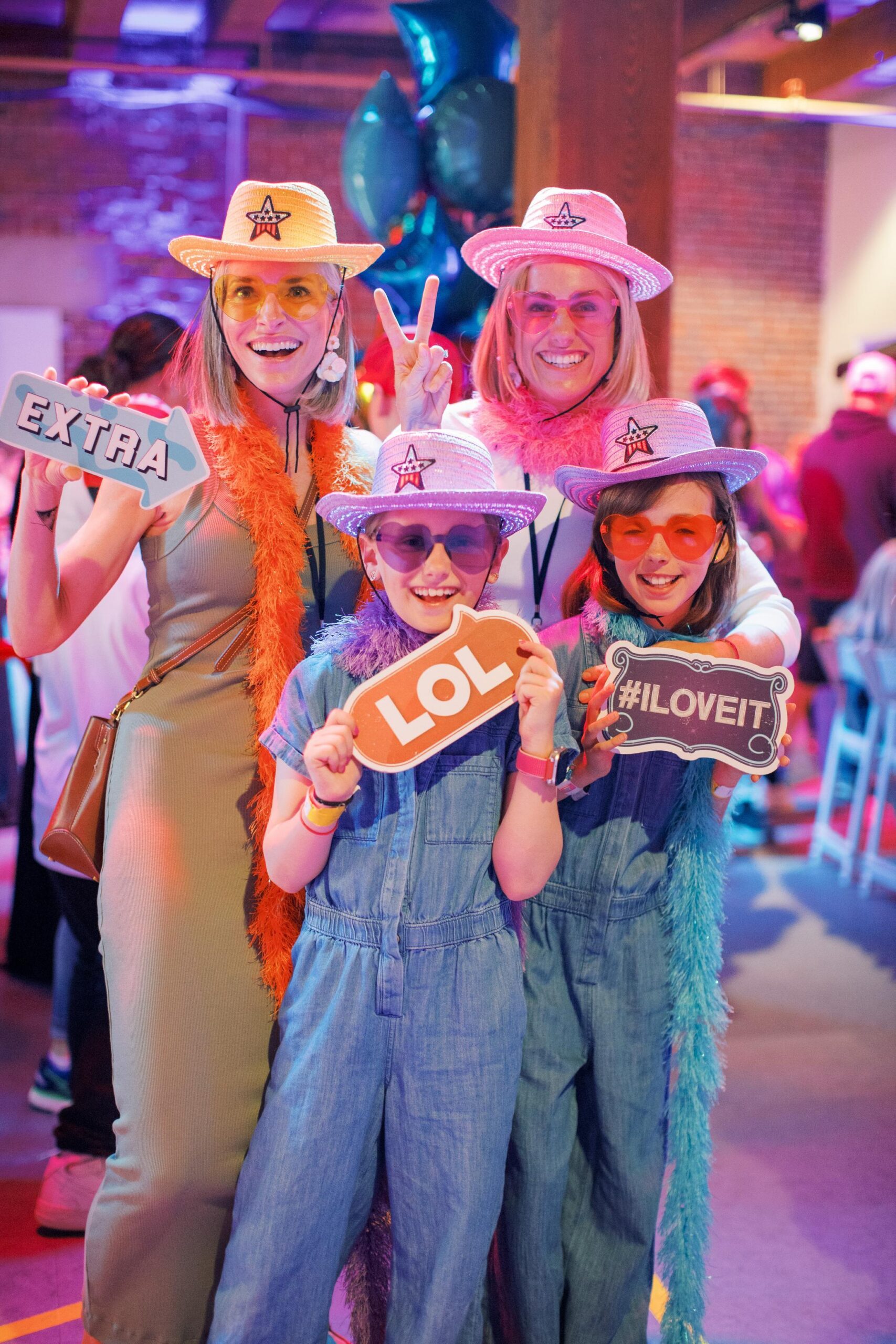 Four people wearing colorful hats, sunglasses, and feather boas pose at a party, holding signs that say EXTRA, LOL, and #ILOVEIT, with smiling faces and a festive atmosphere around them.