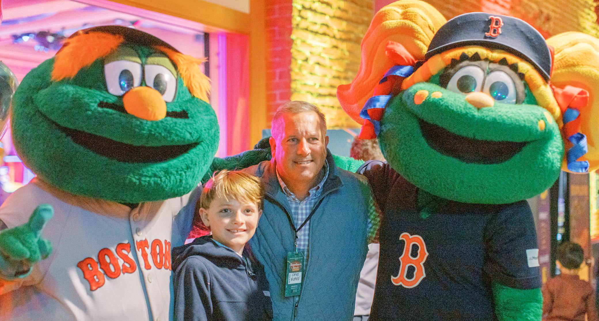 A man and a boy smile for a photo with two people in Boston Red Sox mascot costumes, standing indoors with warm lighting and a brick wall background.