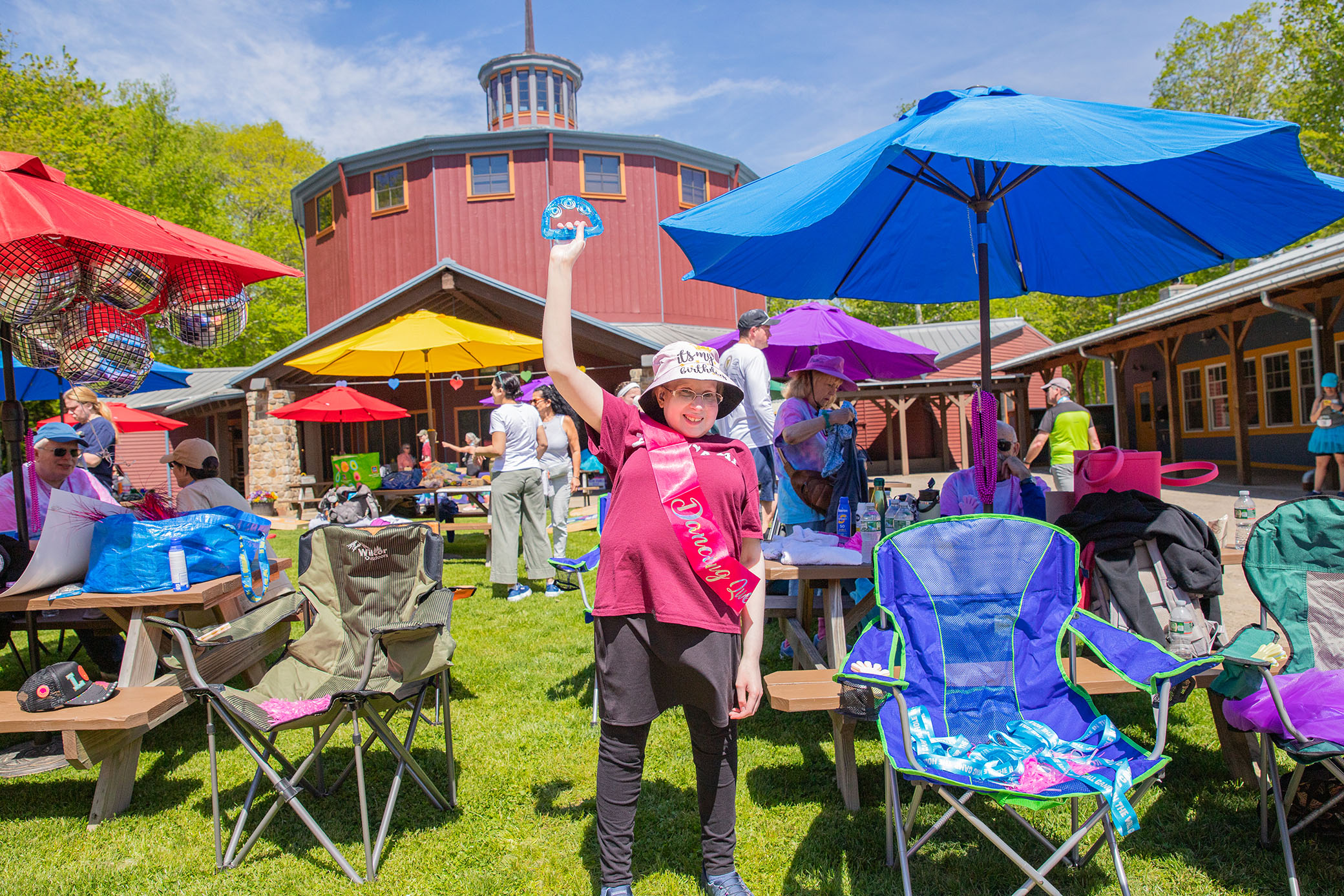 A smiling child in a birthday sash and hat stands on grass, holding up a blue frosted cookie. Colorful umbrellas, chairs, and people fill the bright outdoor picnic area in front of a red building.
