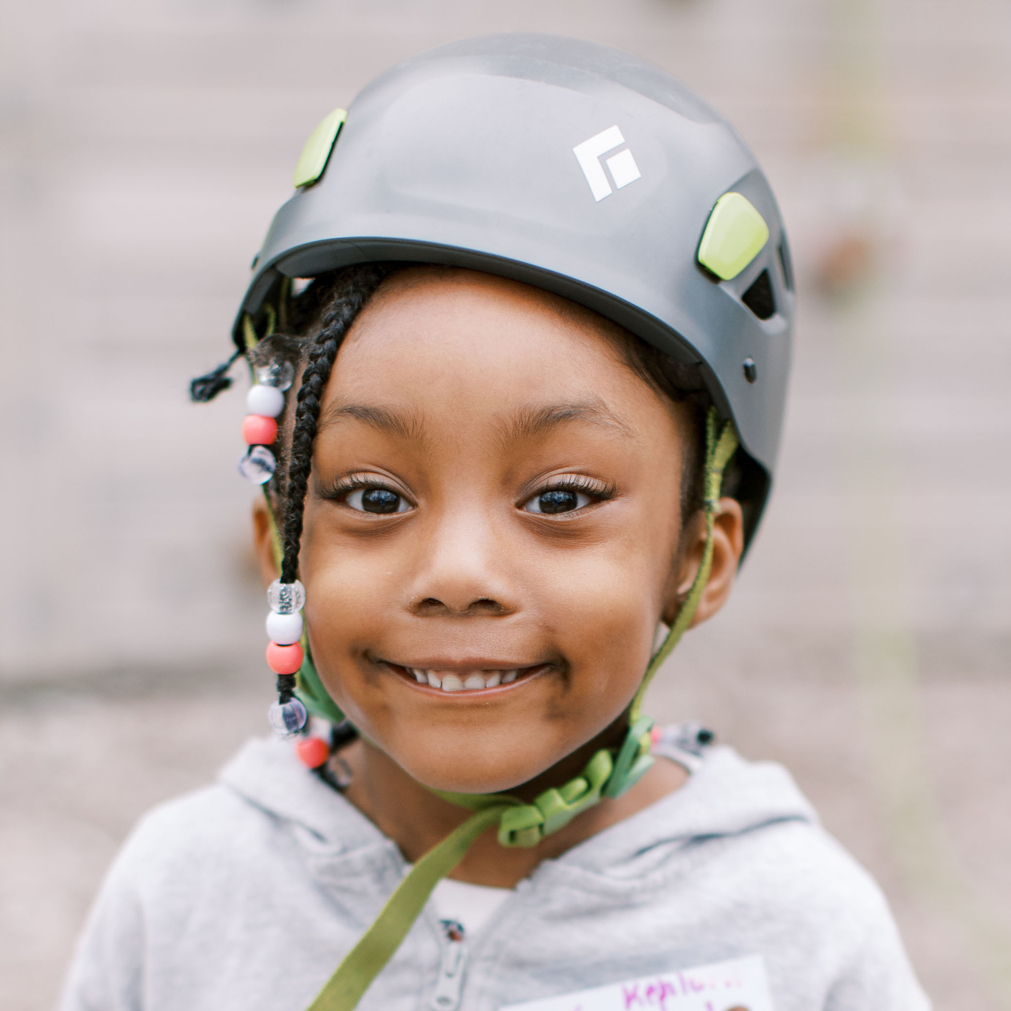 A young child with braids and colorful beads smiles while wearing a gray climbing helmet and a light hoodie, standing outdoors.