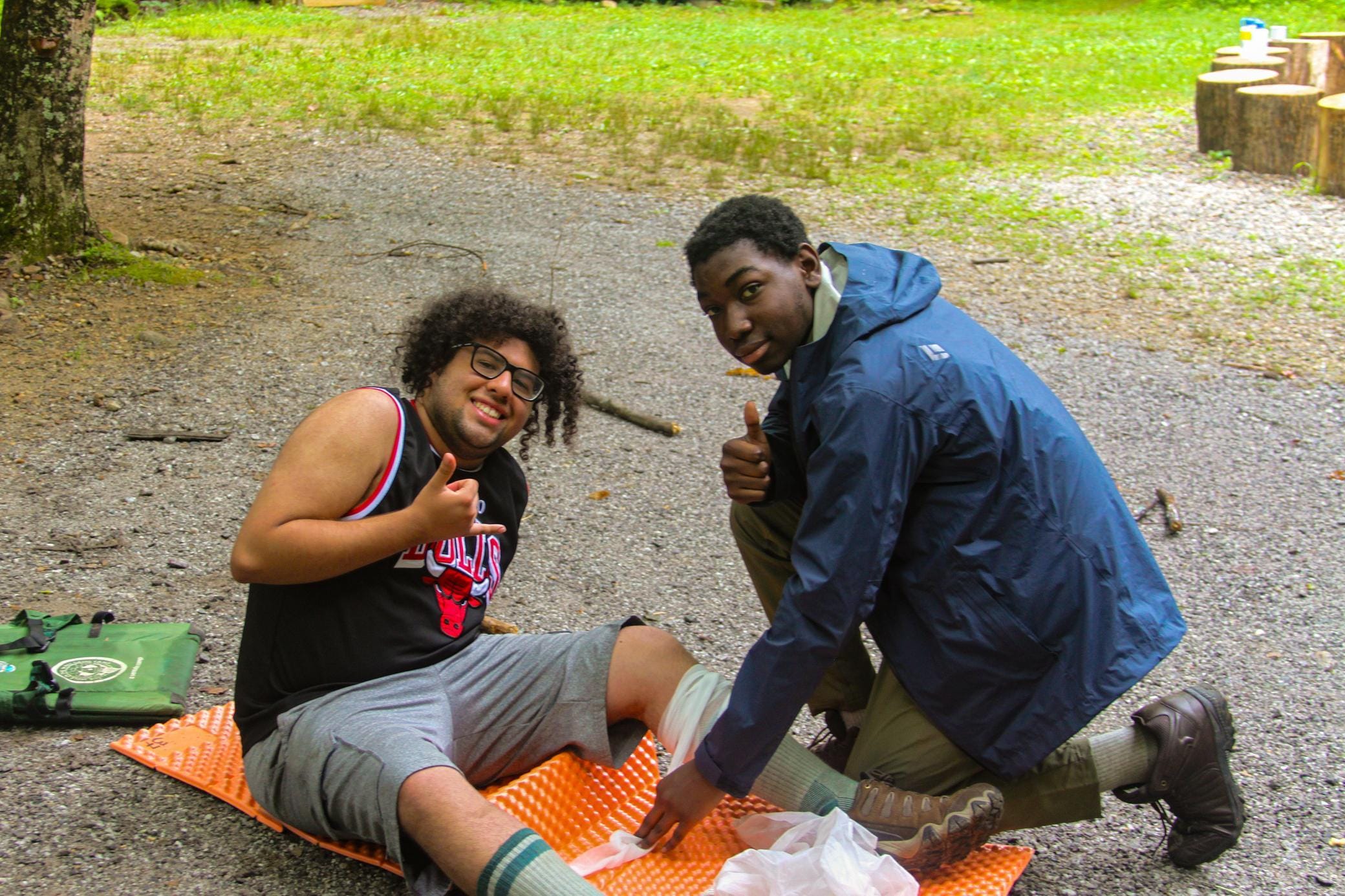 Two people sit outdoors; one has their leg bandaged and is smiling with a thumbs up, while the other, wearing a jacket, kneels next to them, also giving a thumbs up. There is greenery and gravel around them.