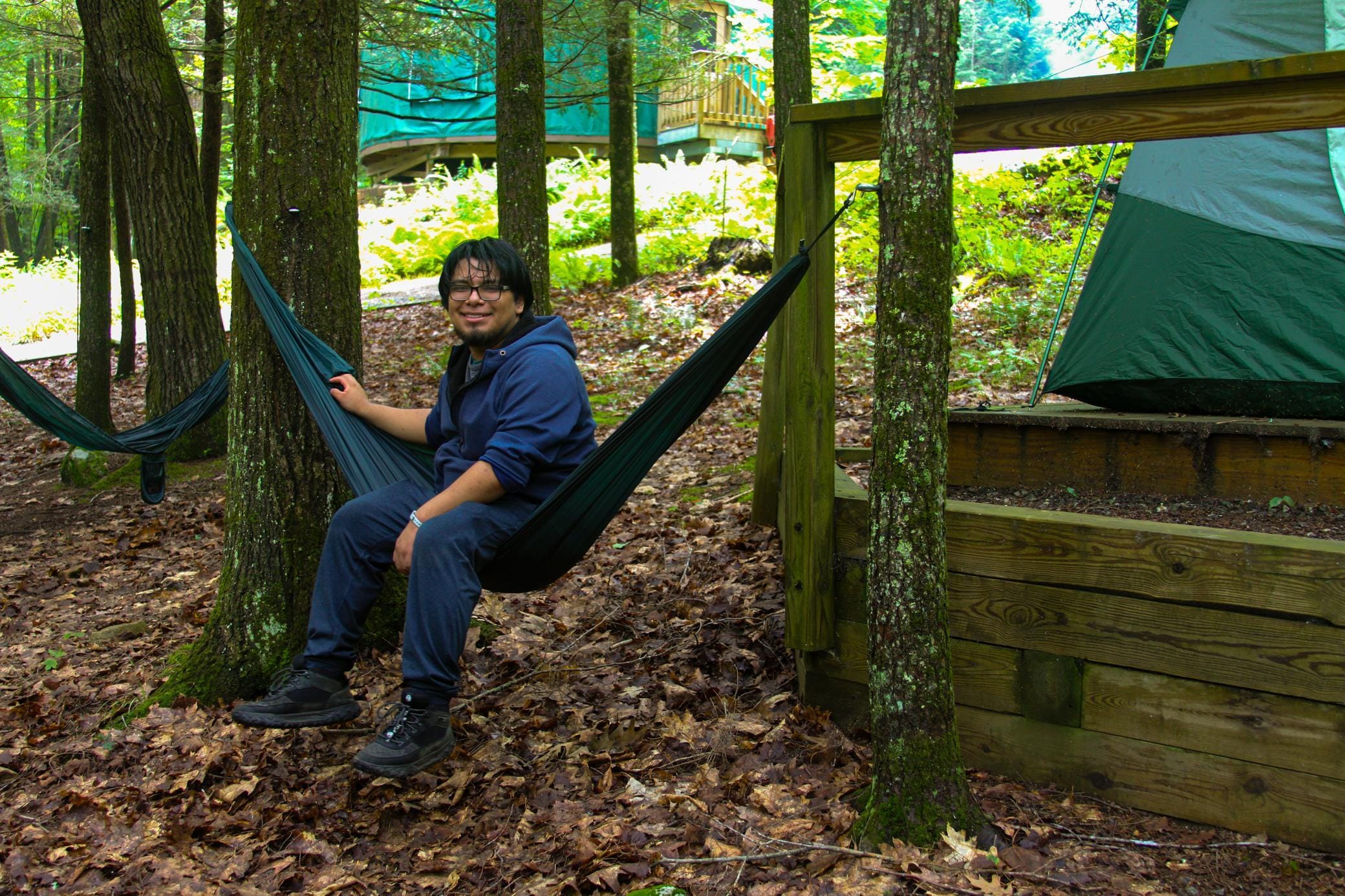 A person in a blue hoodie sits on a hammock between trees in a wooded campsite, with a tent and wooden structure nearby.