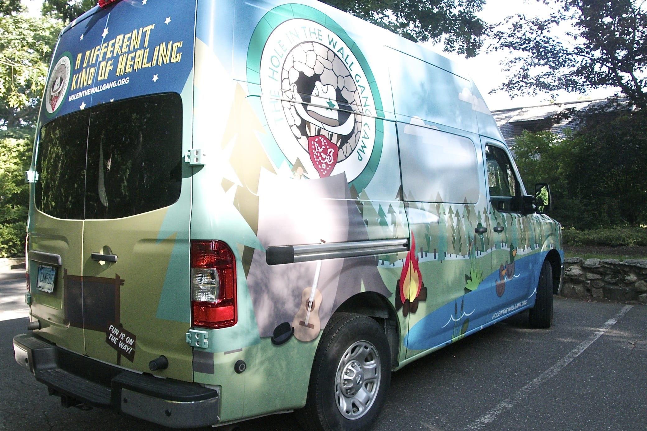 A colorful van with a forest and campfire mural, logo with eagle, and text A Different Kind of Healing is parked outside on a sunny day near trees and a stone wall.