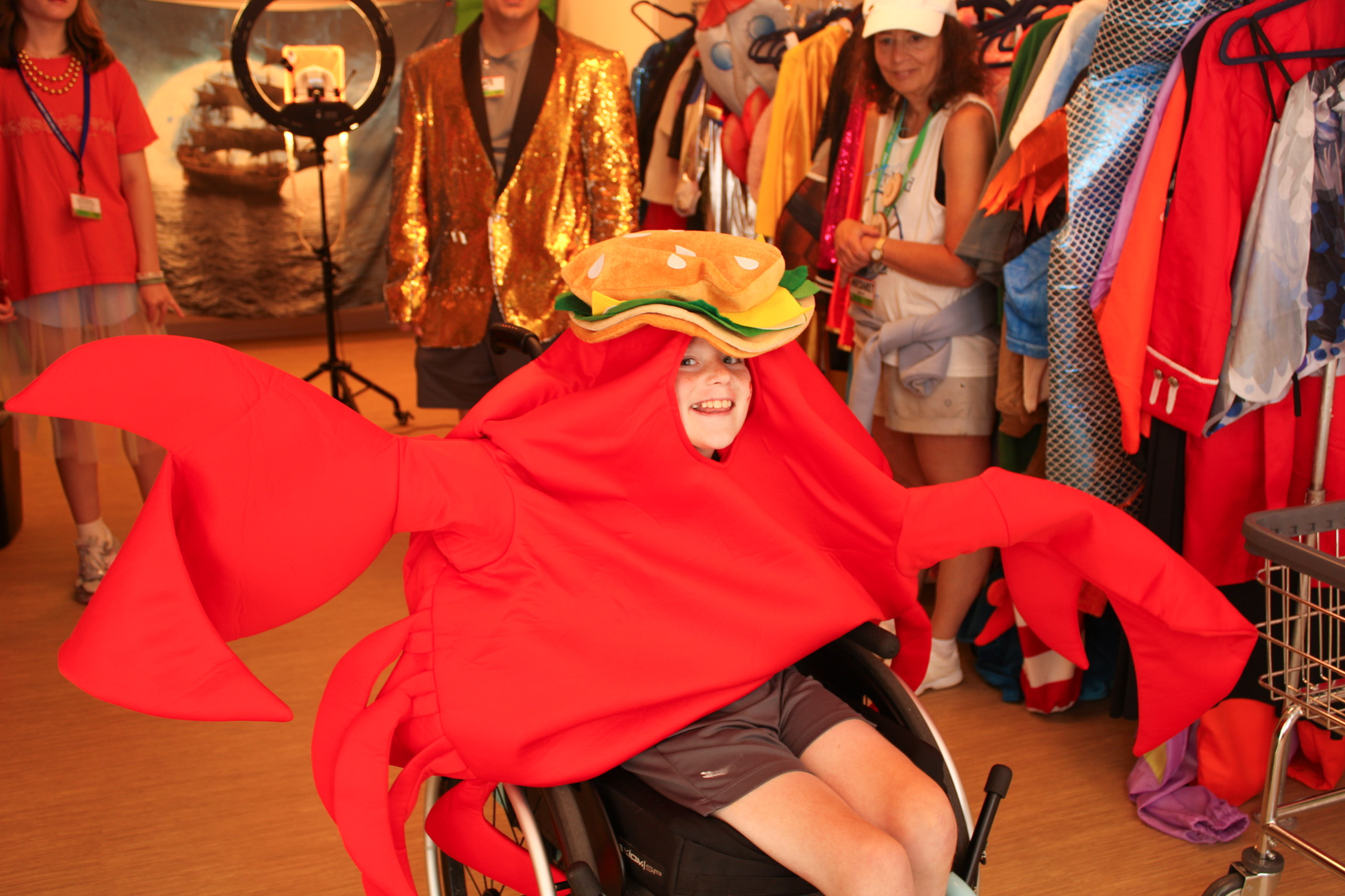 A child in a wheelchair smiles while wearing a bright red crab costume with a burger hat, surrounded by people and colorful costumes in a lively room.