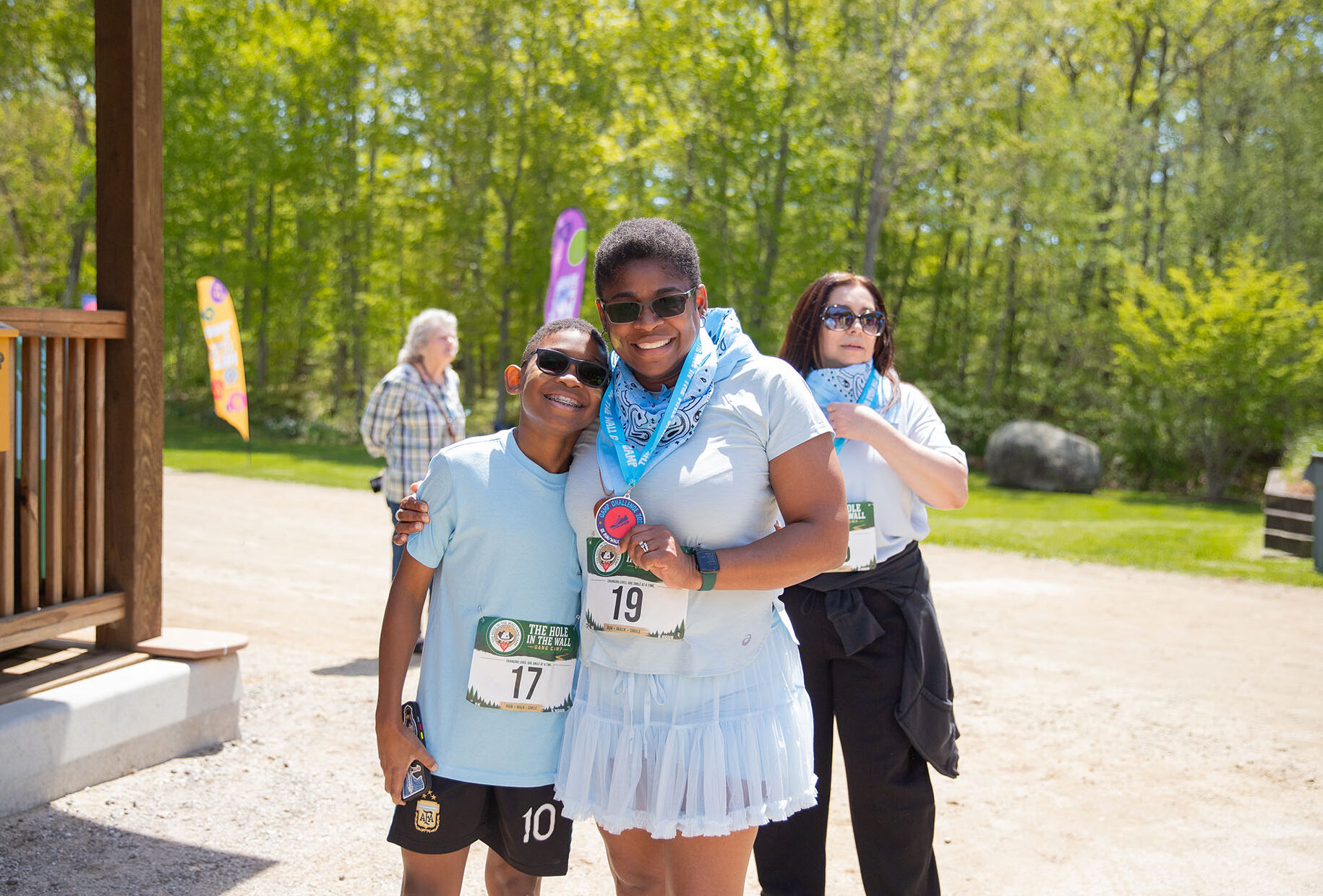 Two people wearing light blue shirts and race bibs smile and pose together outdoors, while a third person stands behind them tying a blue bandana. Lush green trees and bright sunlight fill the background.