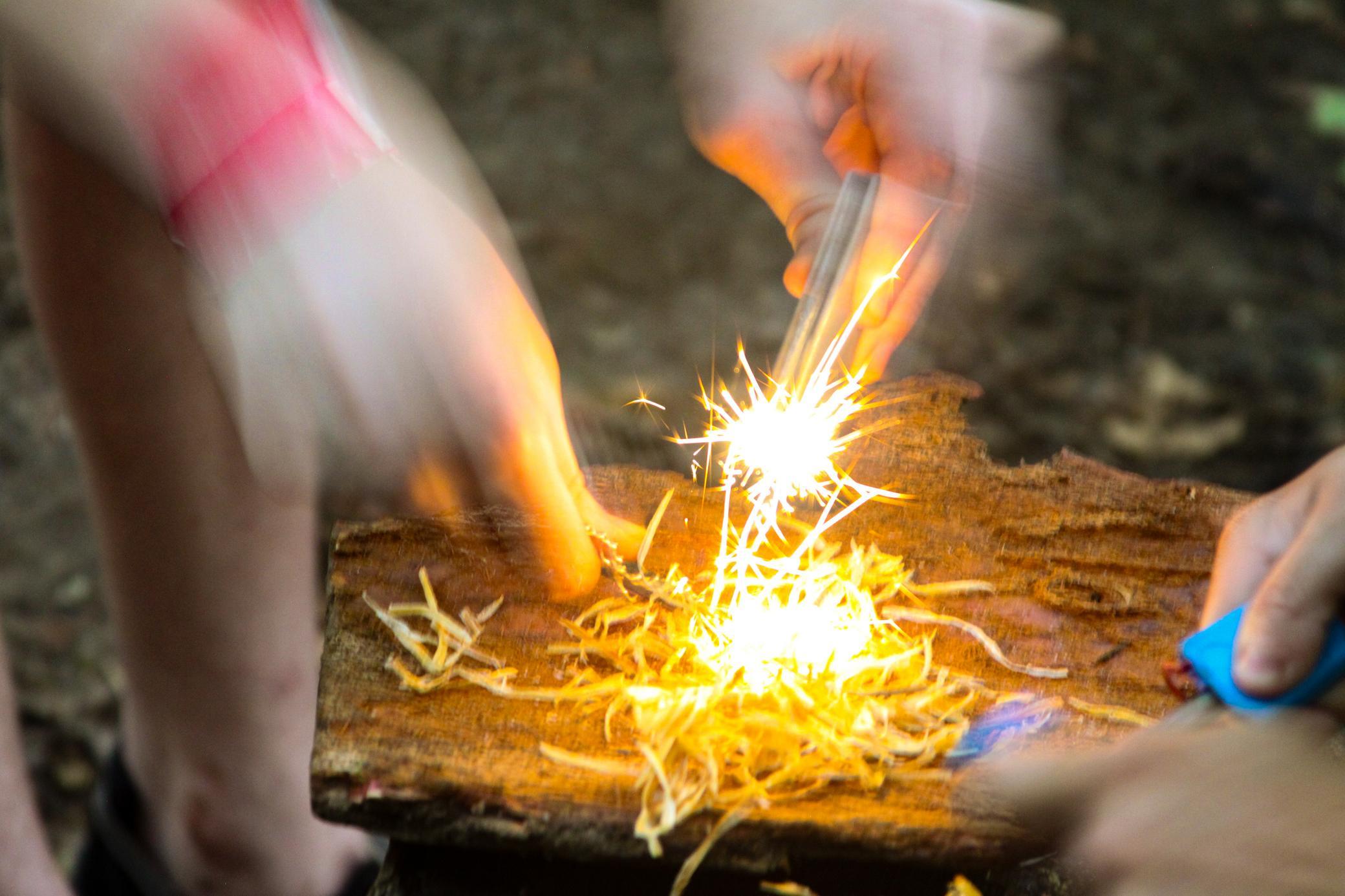 Three hands create sparks with a firestarter above dry kindling on a wooden surface, attempting to start a fire outdoors. The image is dynamic, showing motion blur and bright sparks.