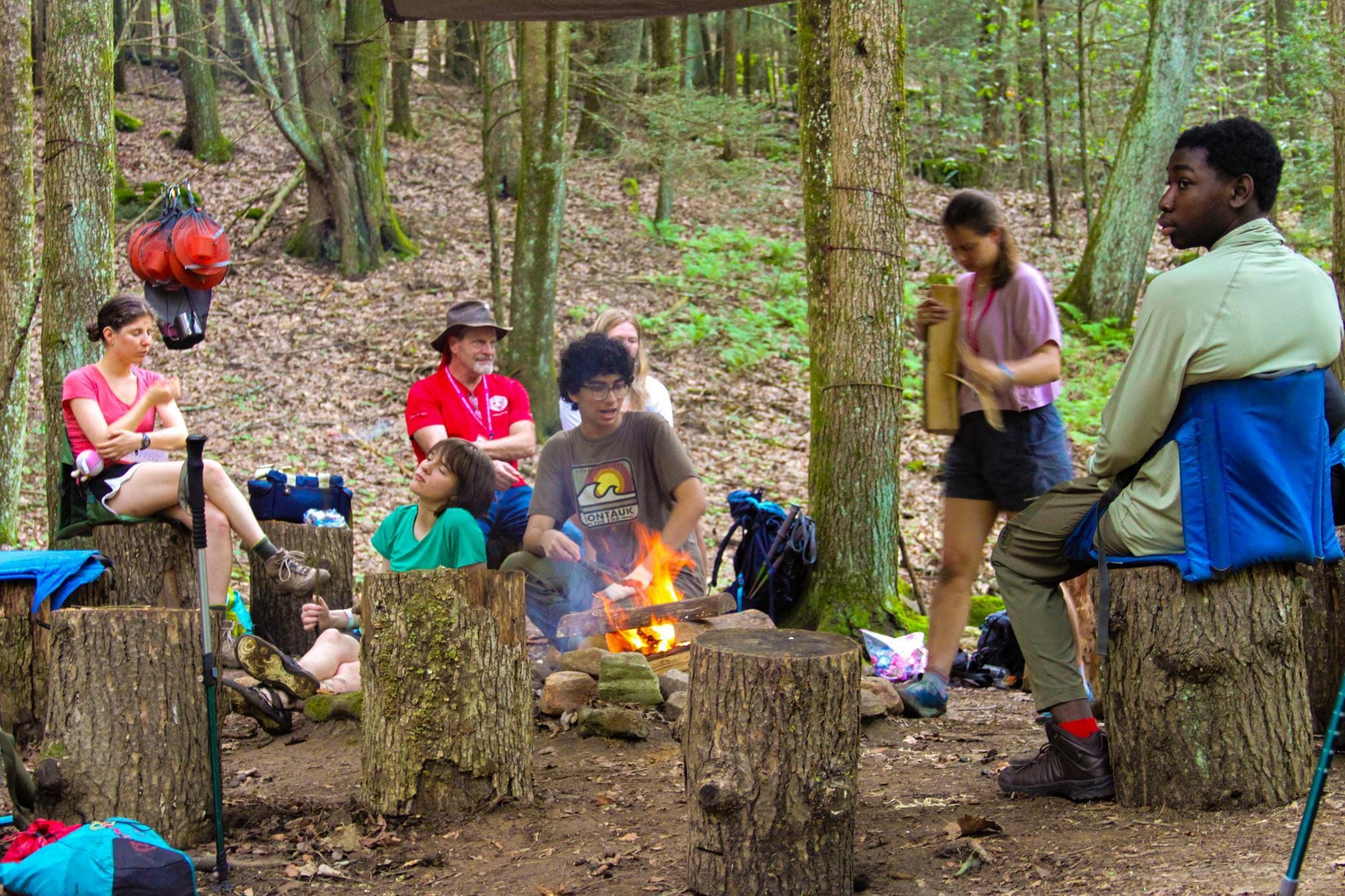 A group of people sits on tree stumps around a campfire in a forest, talking and relaxing. Some are wearing outdoor clothing, and backpacks and gear are visible in the background among the trees.