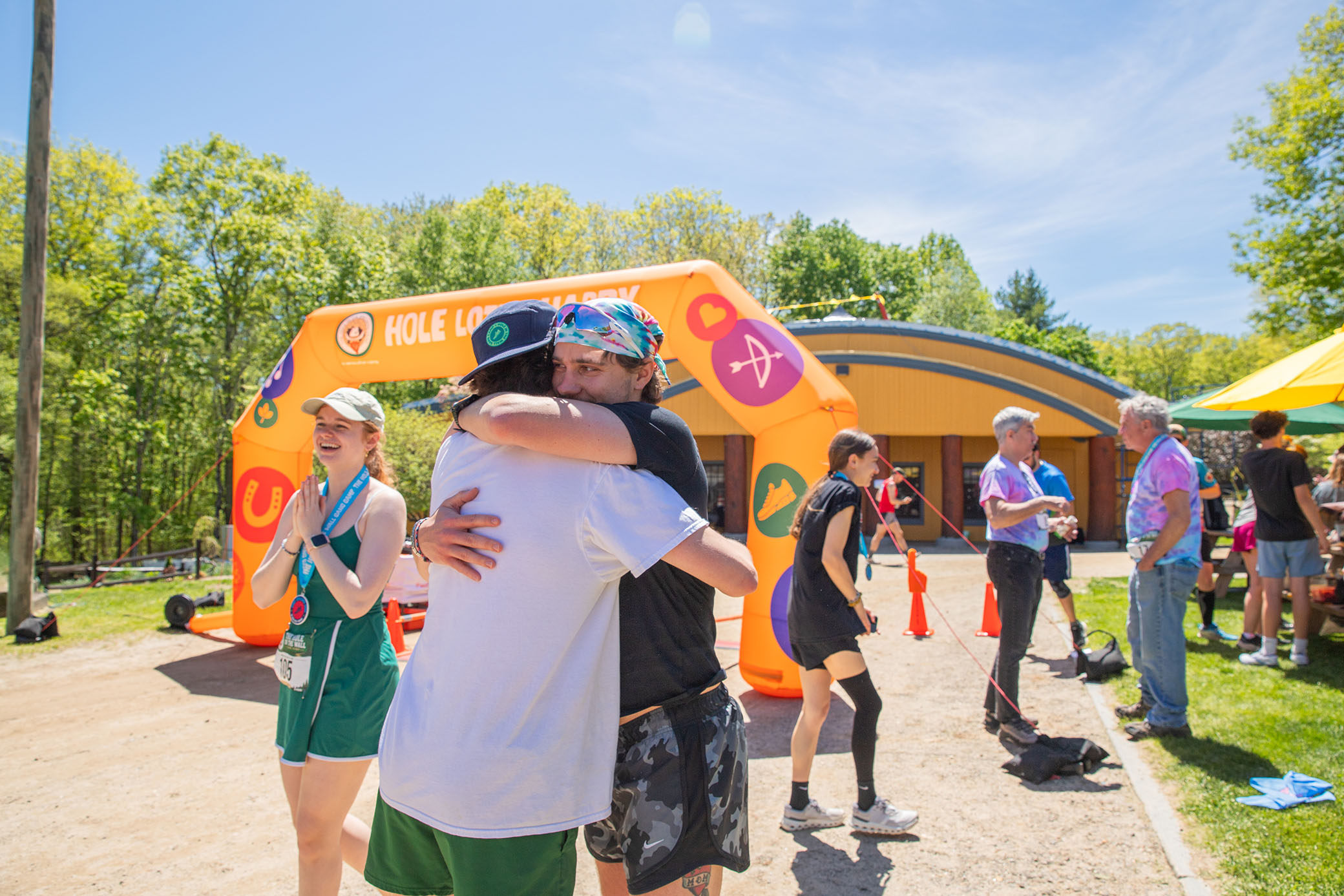 Two people hug near an inflatable arch at an outdoor event. Others stand nearby, some wearing medals and athletic gear. Bright sunlight and green trees suggest a summer day.