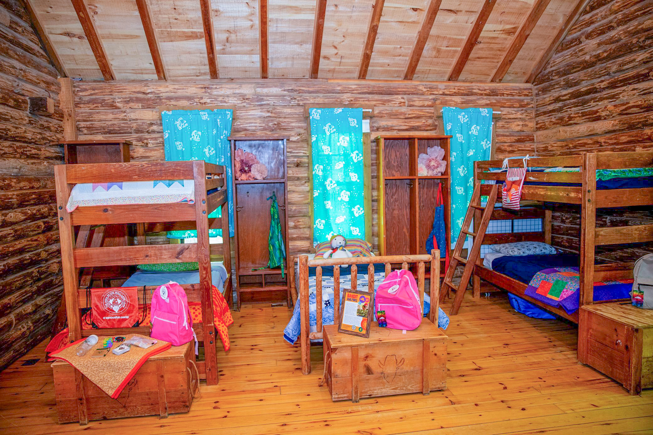 A cozy log cabin room with three wooden bunk beds, colorful quilts, blue curtains, and pink backpacks on a table and the beds. The walls and ceiling are made of natural wood.