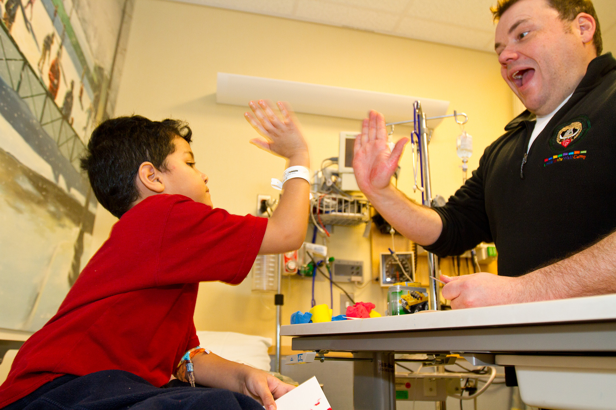 A young boy in a hospital bed and a man sitting nearby are smiling and giving each other a high five in a cheerful hospital room setting.