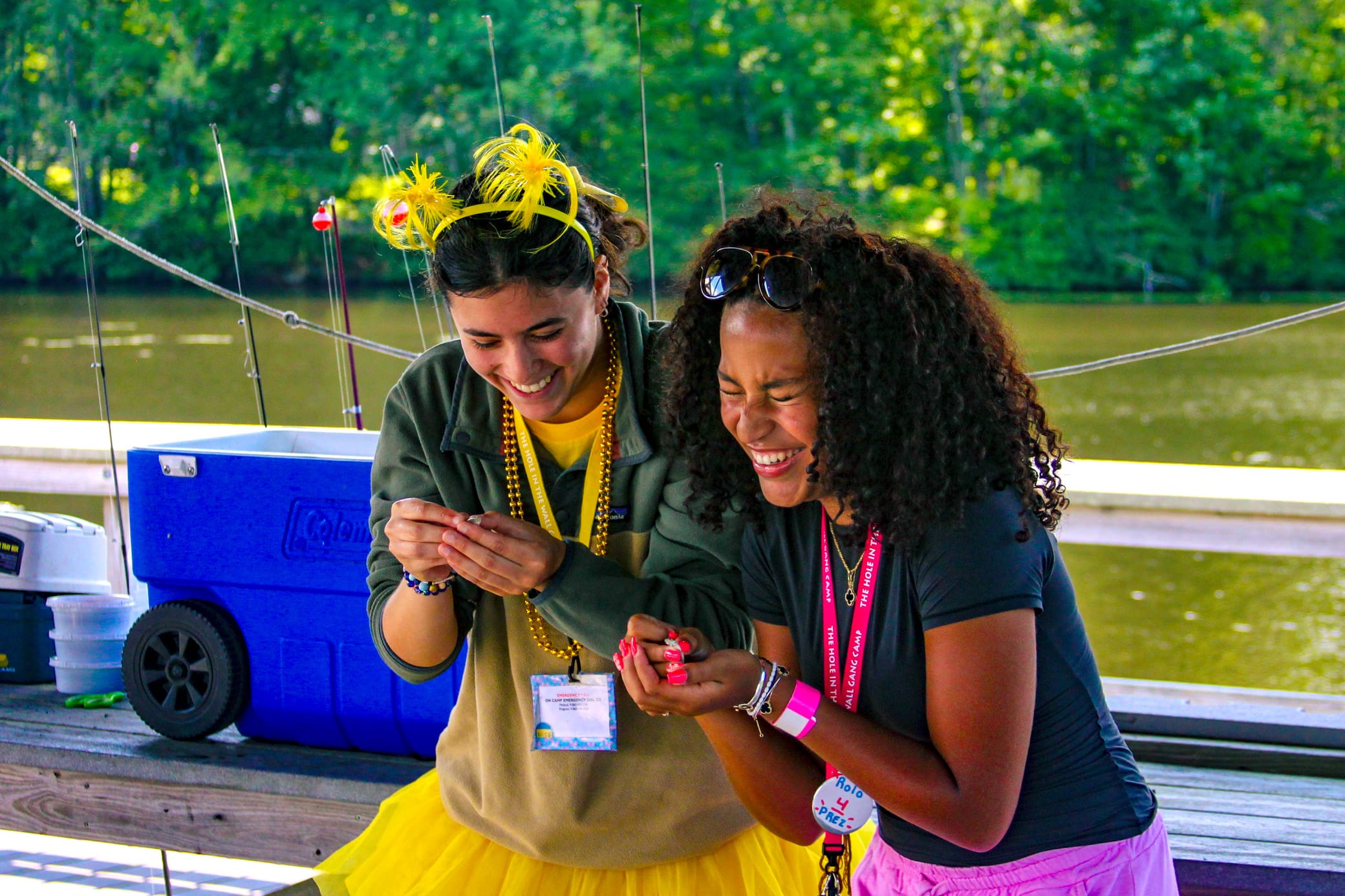 Two women smile and laugh together while fishing by a lake. One wears a yellow tutu, beads, and feather headband. The other wears sunglasses and a pink badge lanyard. A blue cooler and fishing rods are in the background.