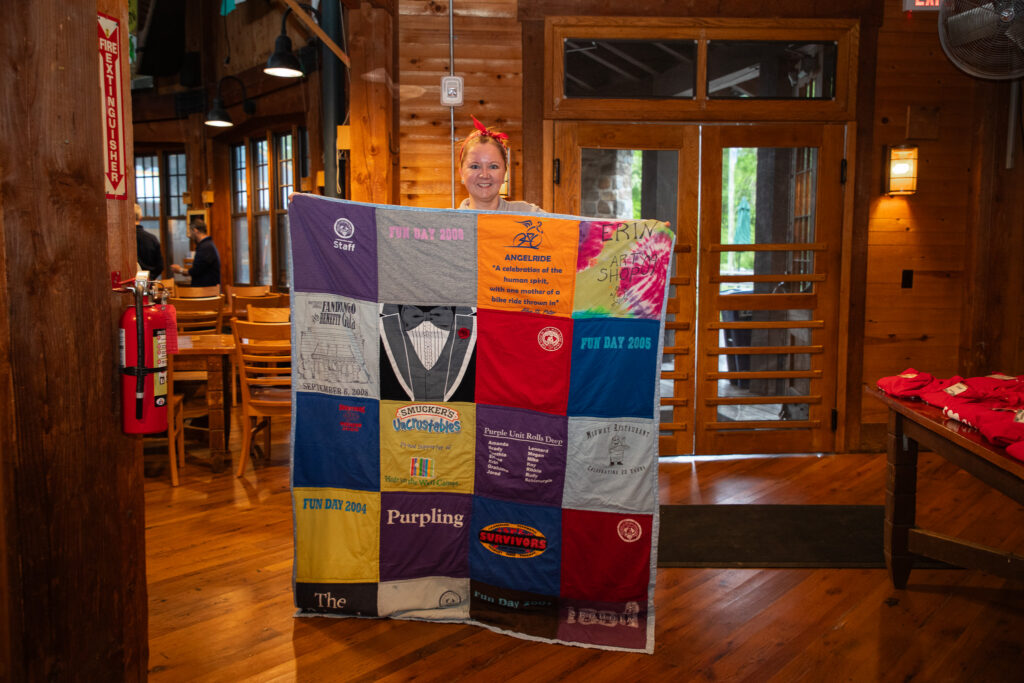 A person stands indoors, smiling and holding up a colorful quilt made from various T-shirts. The room has wooden floors and walls, with tables, chairs, and a visible fire extinguisher in the background.