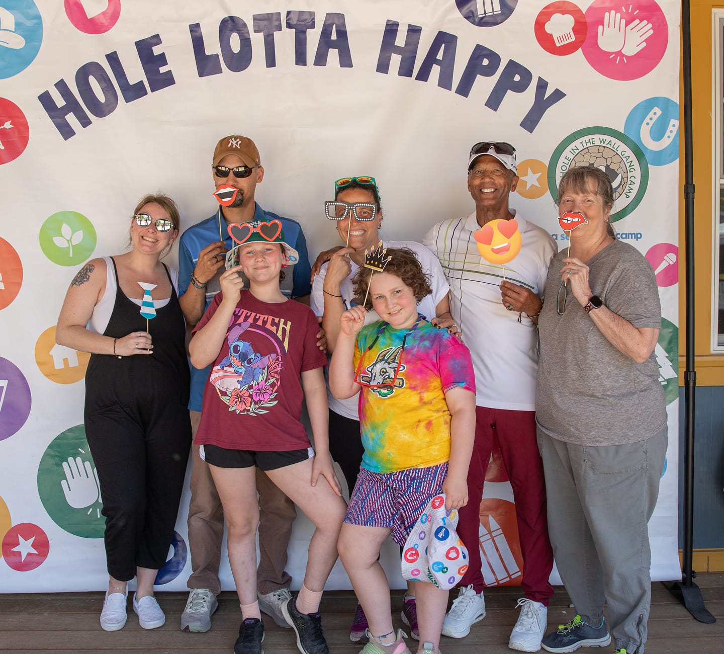 Seven people pose in front of a Hole Lotta Happy banner, smiling and holding fun photo props like heart glasses, crowns, and mustaches. The group looks cheerful and playful, dressed casually for a festive event.