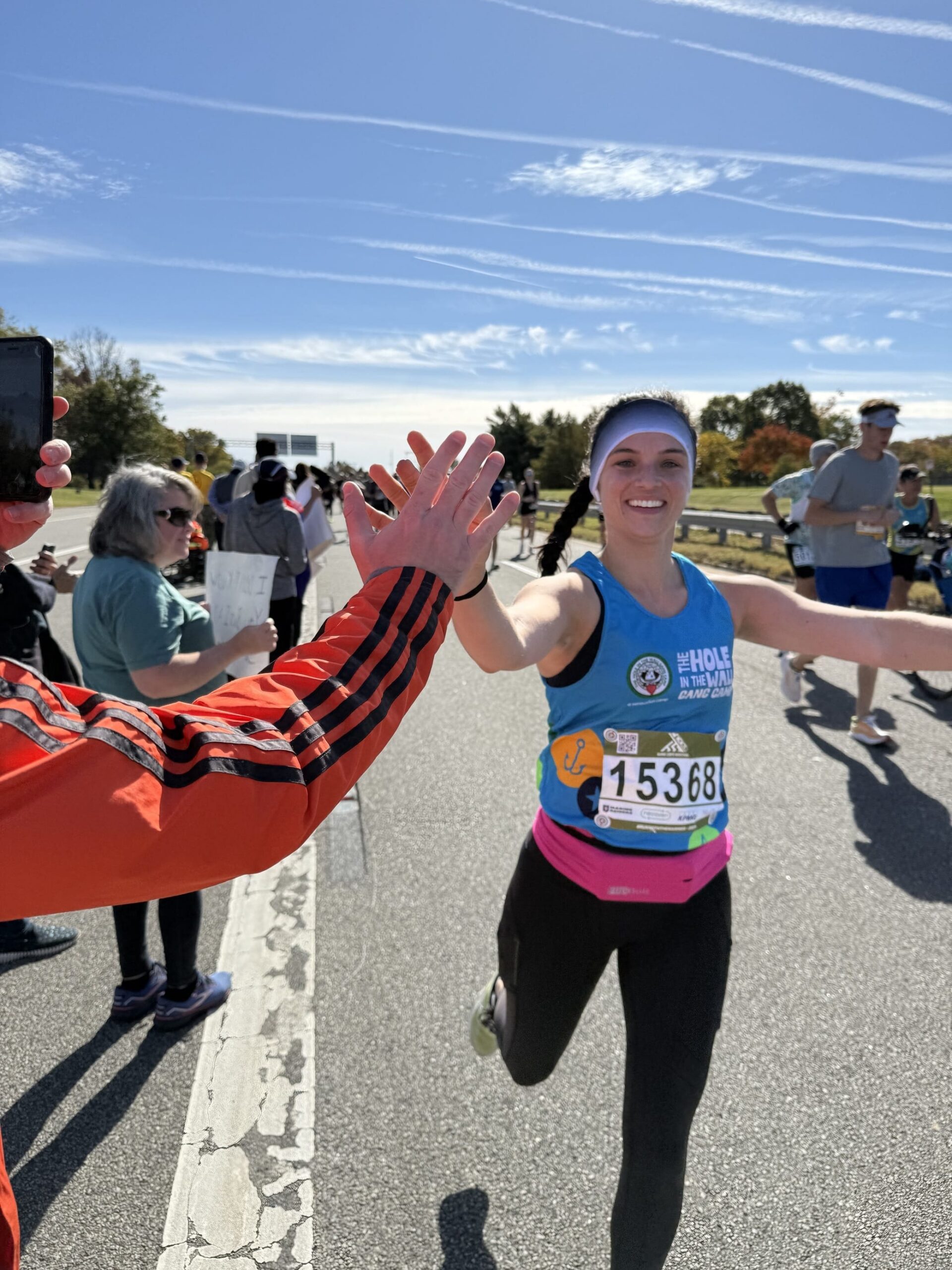 A smiling runner in a blue tank top, wearing race bib 15368, gives a high five to a supporter. Other spectators stand nearby along the sunny road, under a blue sky with streaks of clouds.