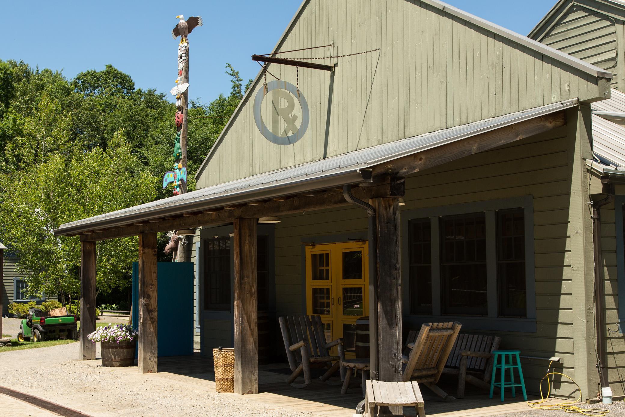 A rustic wooden building with a porch, rocking chairs, yellow double doors, and a totem pole outside, set on a sunny day with trees in the background.