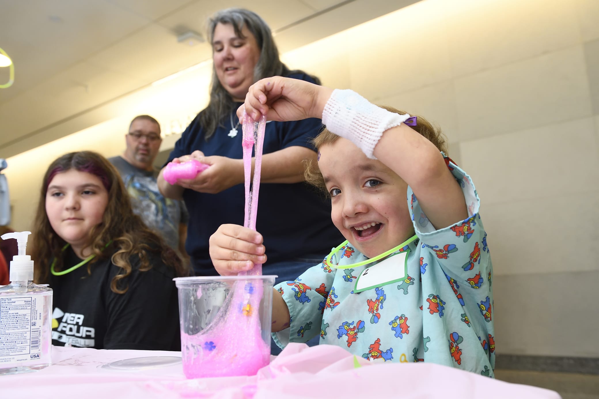 A young child in a hospital gown smiles while playing with pink slime at a table, surrounded by adults and another child. The child has a medical bandage on their arm.