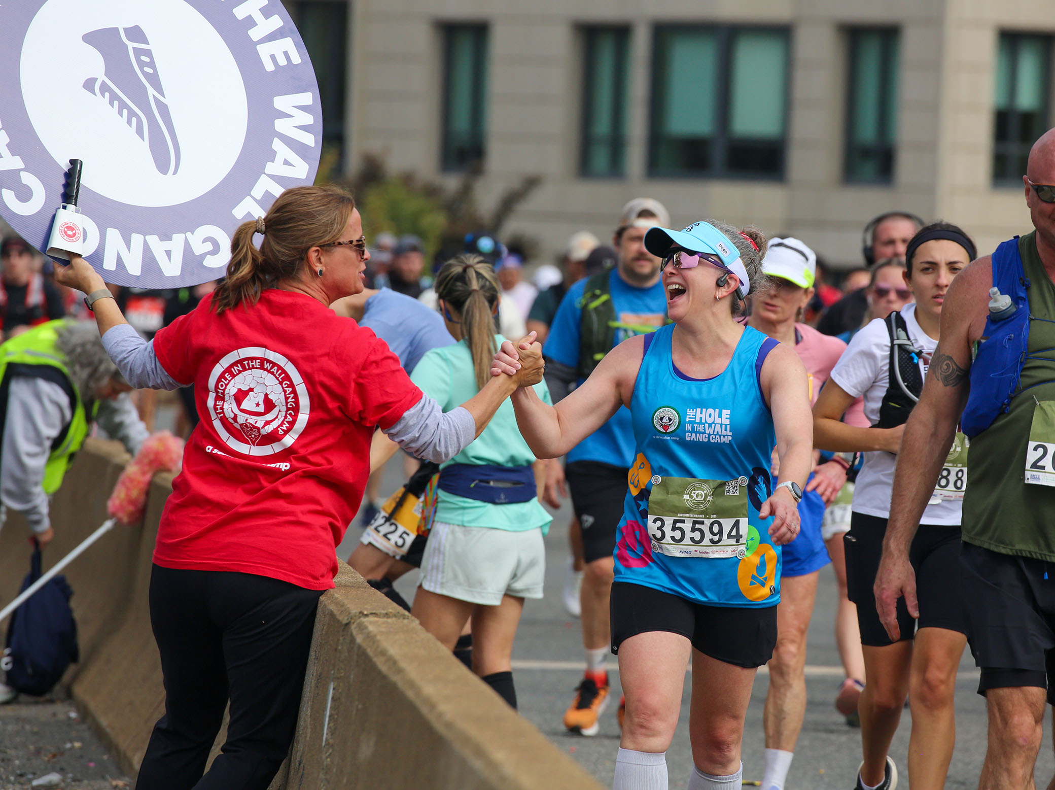 A woman in a red shirt gives a high five to a smiling marathon runner wearing sunglasses and a blue tank top, while other runners continue behind them on a city street.