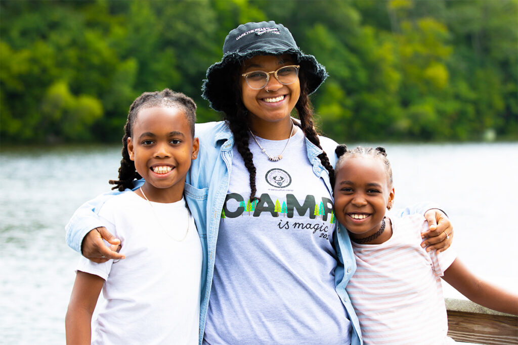 Three smiling children pose together outdoors by a body of water, with green trees in the background. The center child wears glasses, a bucket hat, and a t-shirt that says CAMP is magic..