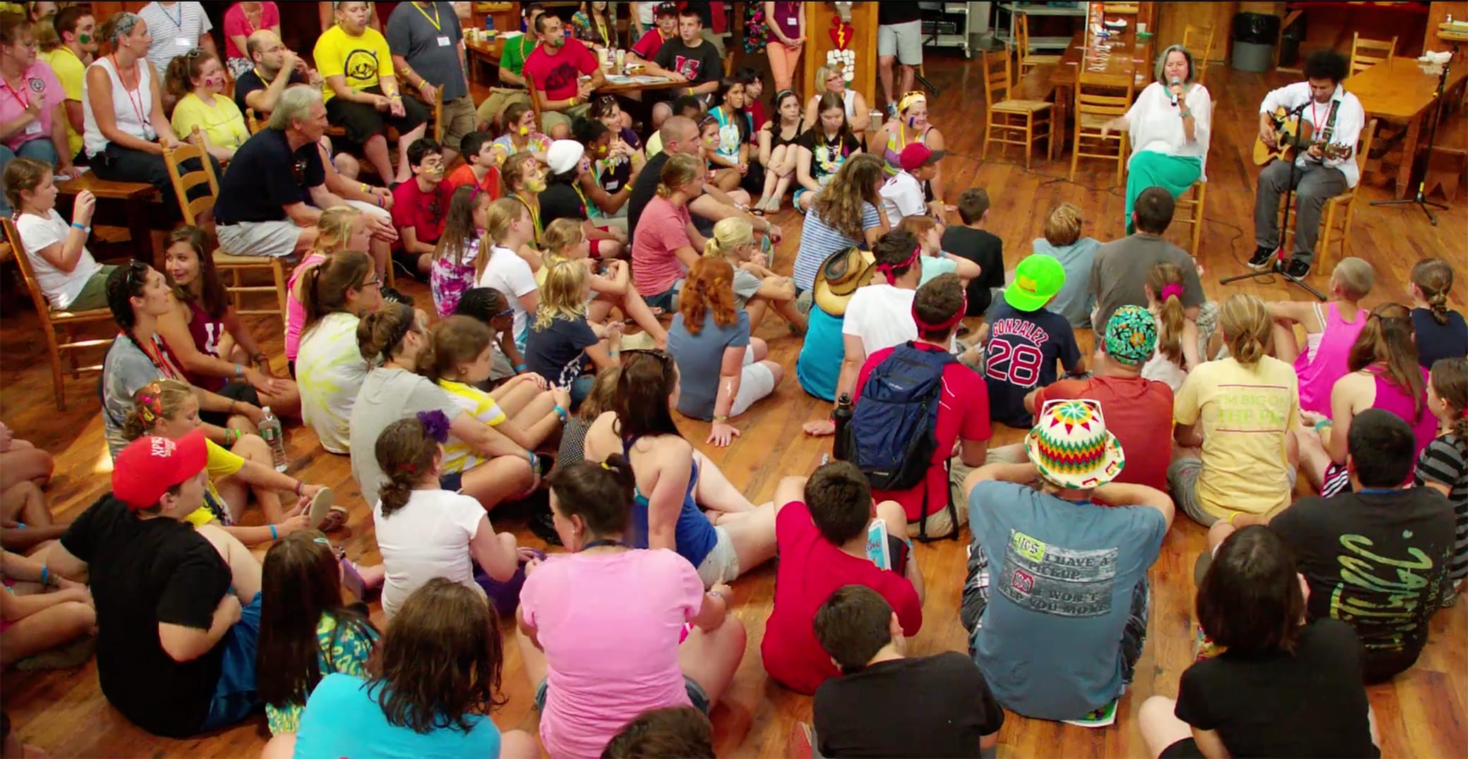 A large group of children sit on a wooden floor in a circle, attentively listening to two people playing guitar and singing at the front of the room.
