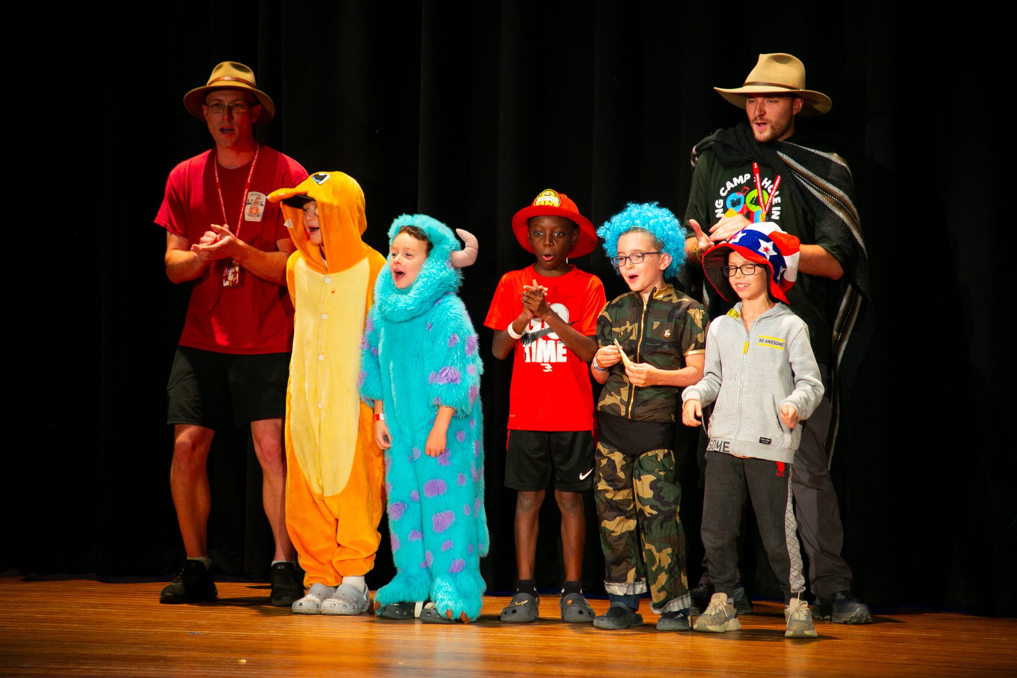 Six people, including children in colorful costumes and two adults in hats, stand on a stage with a black curtain background, appearing to sing or perform together.