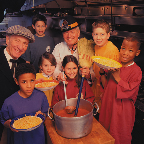 Group of children and two adults in a kitchen, smiling. The children hold plates of spaghetti, and the adults stand behind a pot of tomato sauce, one holding a broom. The scene appears warm and friendly.