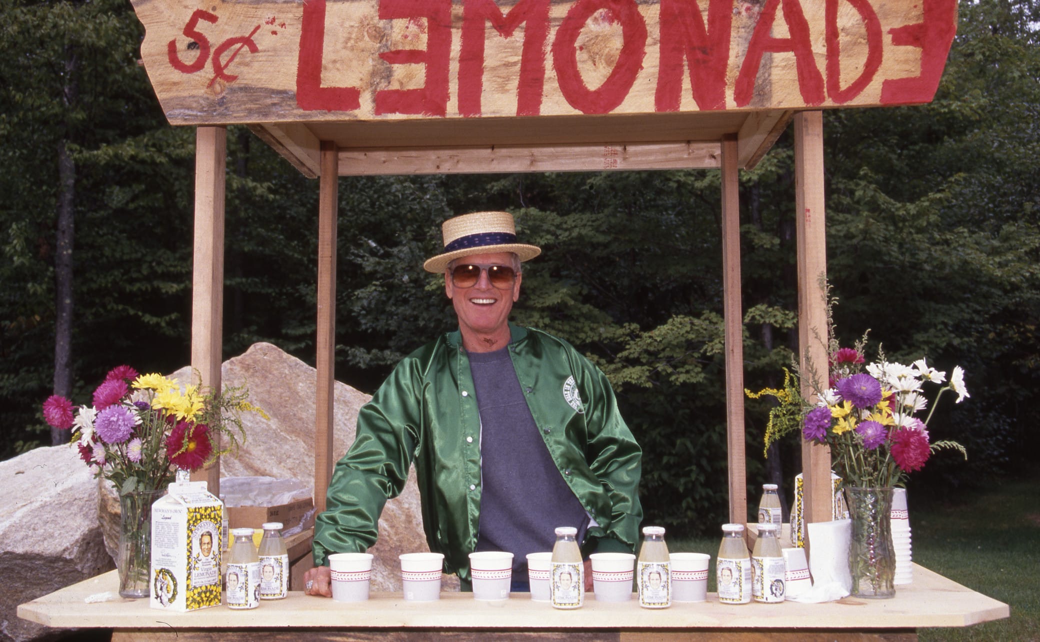 A smiling person in sunglasses and a green jacket stands at a wooden lemonade stand with flowers, cups, and bottles. A sign above reads LEMONADE 5¢ in red letters. Trees and rocks are in the background.
