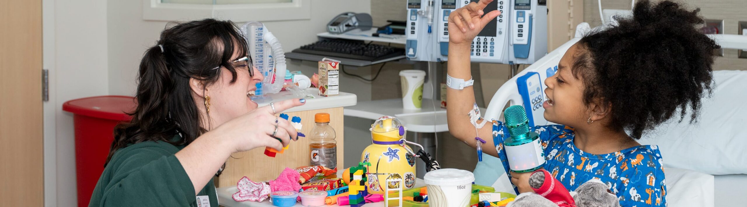 A woman and a young girl in a hospital room laugh and play with colorful toys at a table. The girl is wearing a hospital gown and has medical tubes attached.