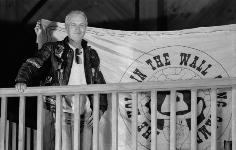 A man in a leather jacket stands on a wooden balcony, smiling, with one hand on the railing and a large banner displayed behind him. A microphone is attached to the railing in front of him.