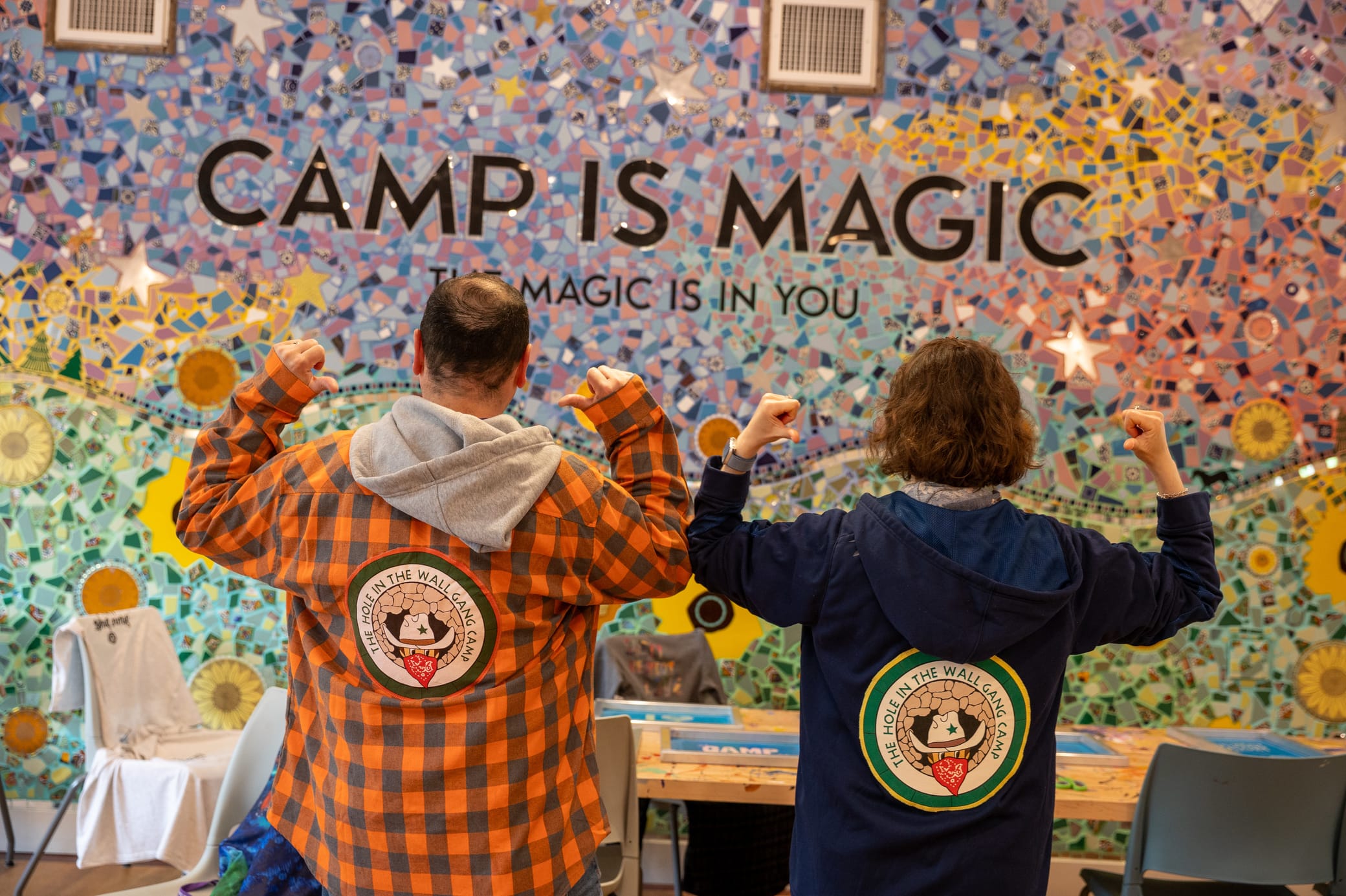 Two people stand with backs to the camera, flexing arms, wearing jackets with matching camp patches. Behind them is a colorful mosaic wall with the words CAMP IS MAGIC and MAGIC IS IN YOU displayed.