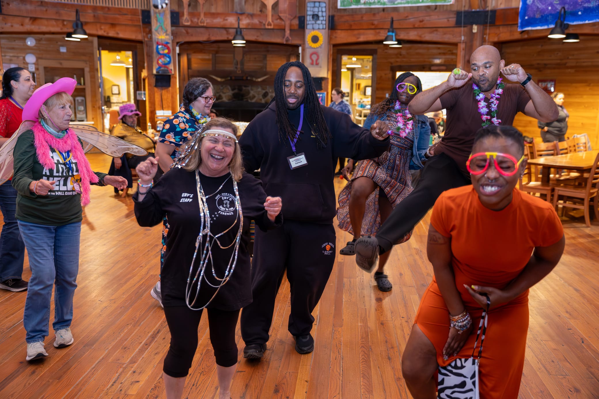 A group of adults in colorful clothes and accessories dance and laugh together in a wooden lodge, enjoying a lively indoor party.