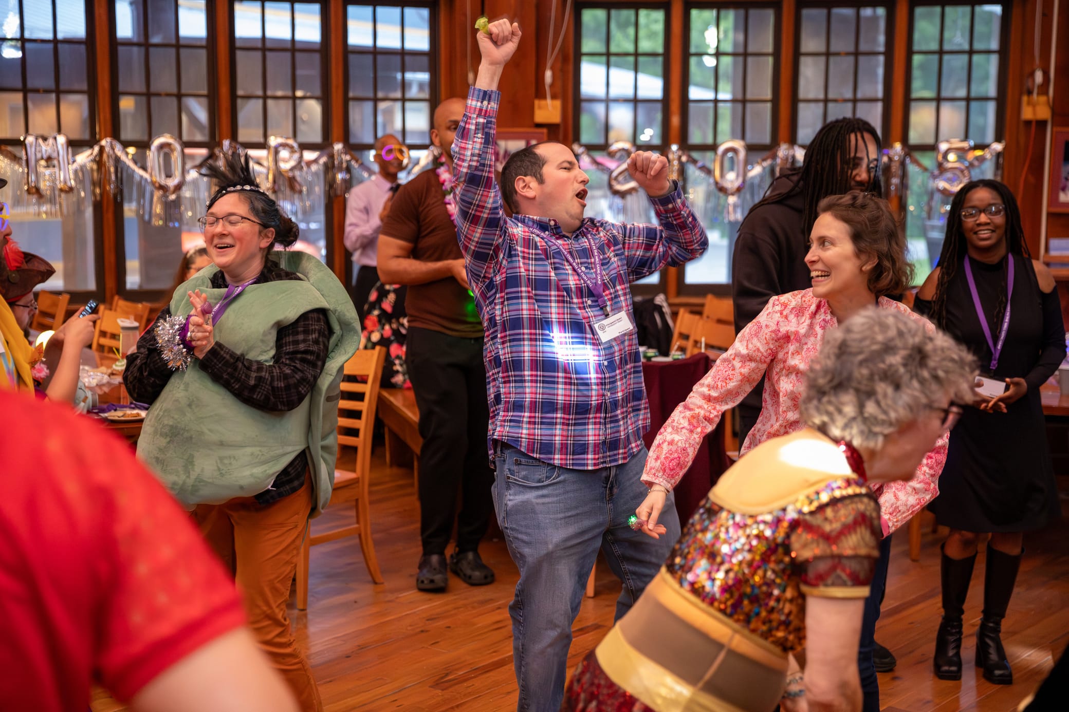 A group of adults in colorful costumes dance and celebrate enthusiastically in a decorated room with wooden floors and large windows.