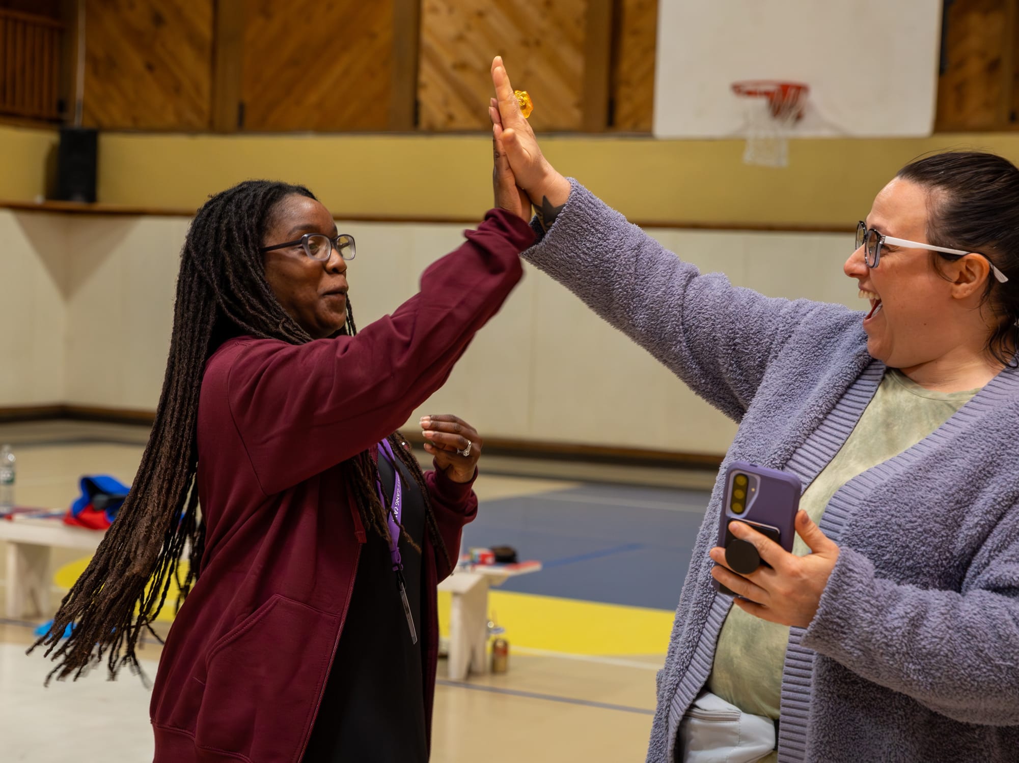 Two women smile and give each other a high five in a gymnasium. One holds a phone and wears glasses, while the other has long braided hair. Benches and a basketball hoop are visible in the background.