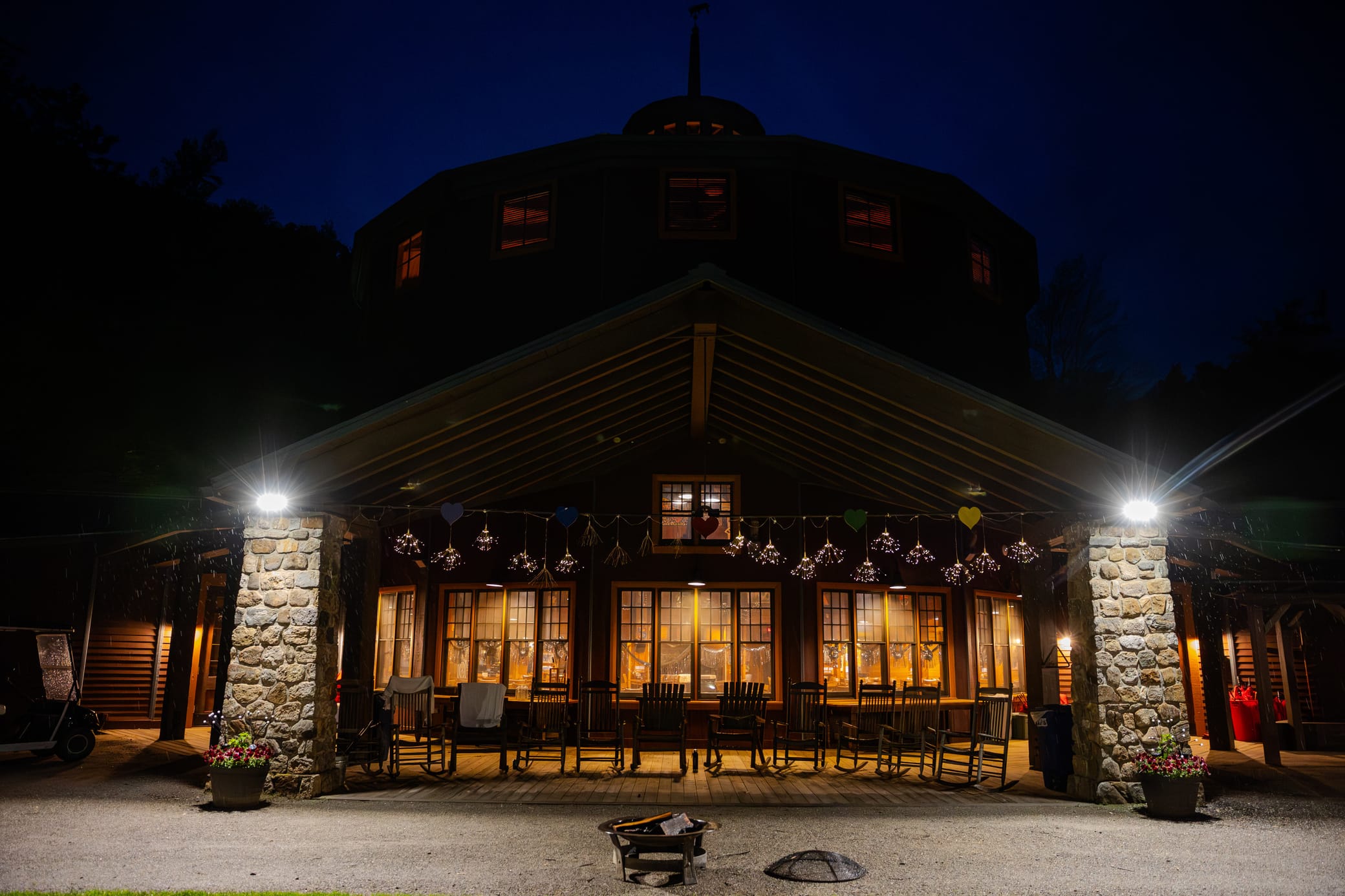 A warmly lit lodge with a stone porch and rocking chairs is seen at night. Overhead lights and string lights decorate the entrance, while a firepit sits unlit in the foreground.