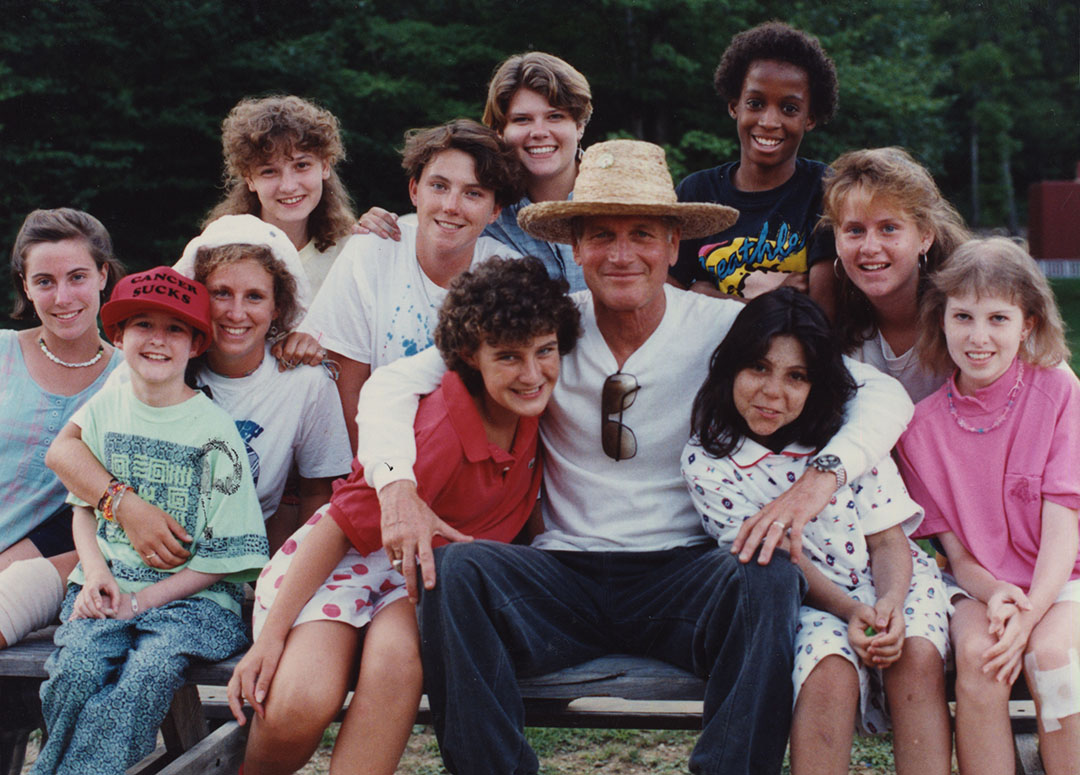 A group of smiling kids and one adult in a straw hat pose together outdoors on a bench, surrounded by trees and greenery.