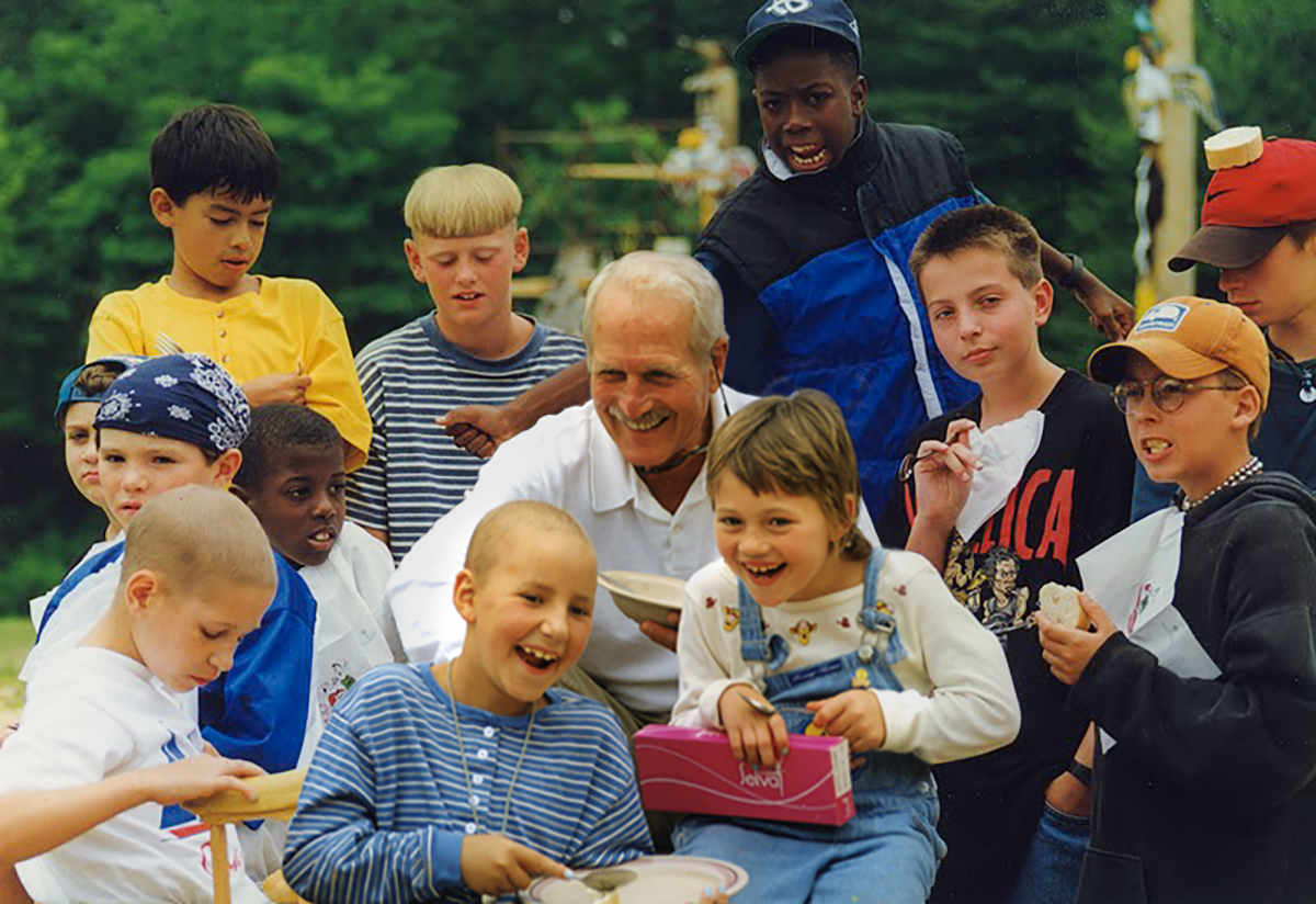 A group of kids and an adult gather outdoors, laughing and smiling, some holding papers and boxes, with trees blurred in the background.