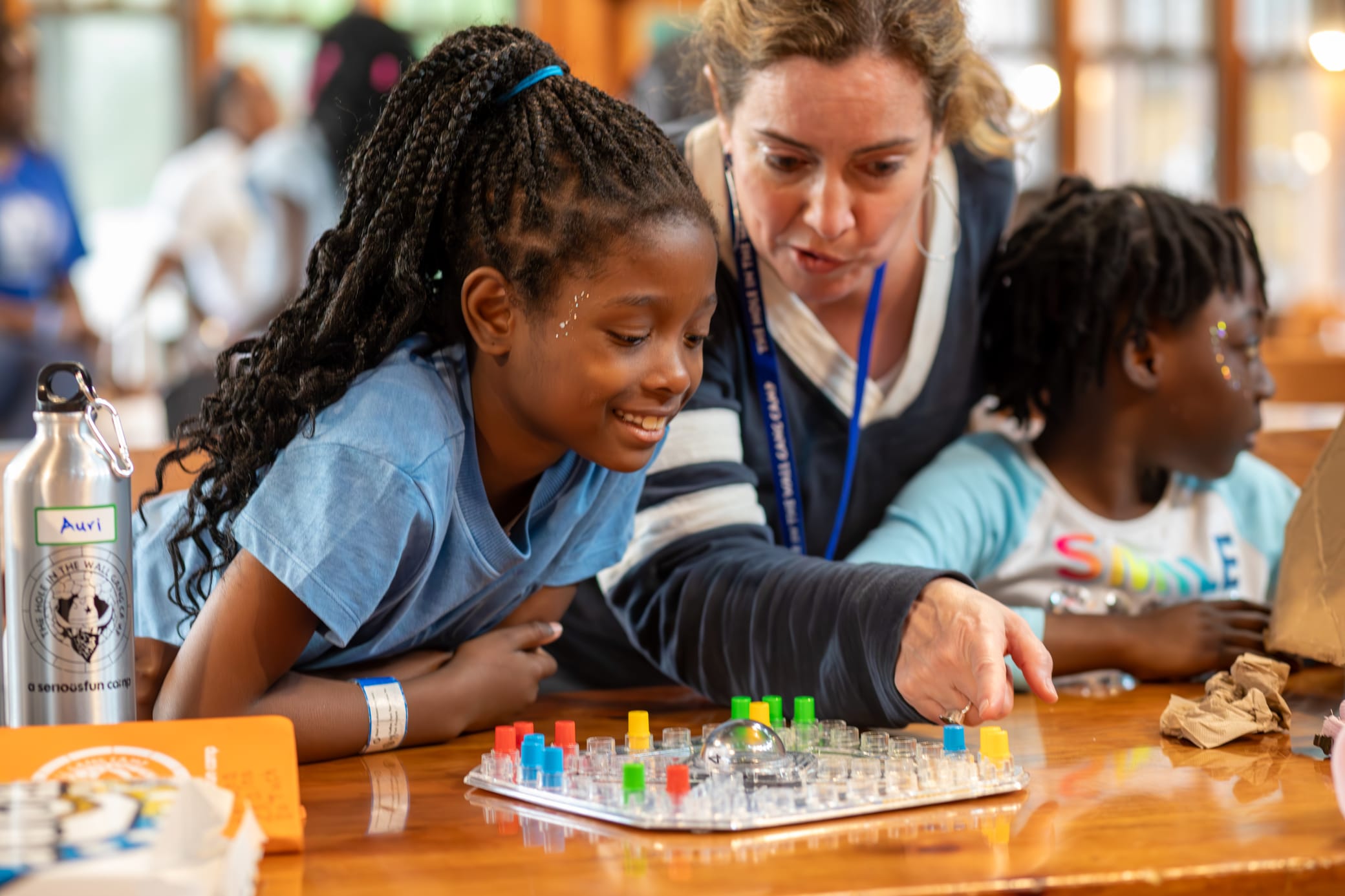 A woman and two girls play a board game at a table. The woman points at the game as the smiling girl watches, while another girl, who faces away, sits beside them. They appear to be enjoying their time together.