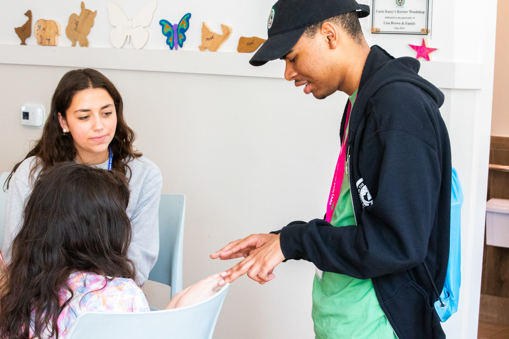 A young man points at a girls hand while she sits at a table, with another young woman watching. Art and decorations are visible on the wall behind them.