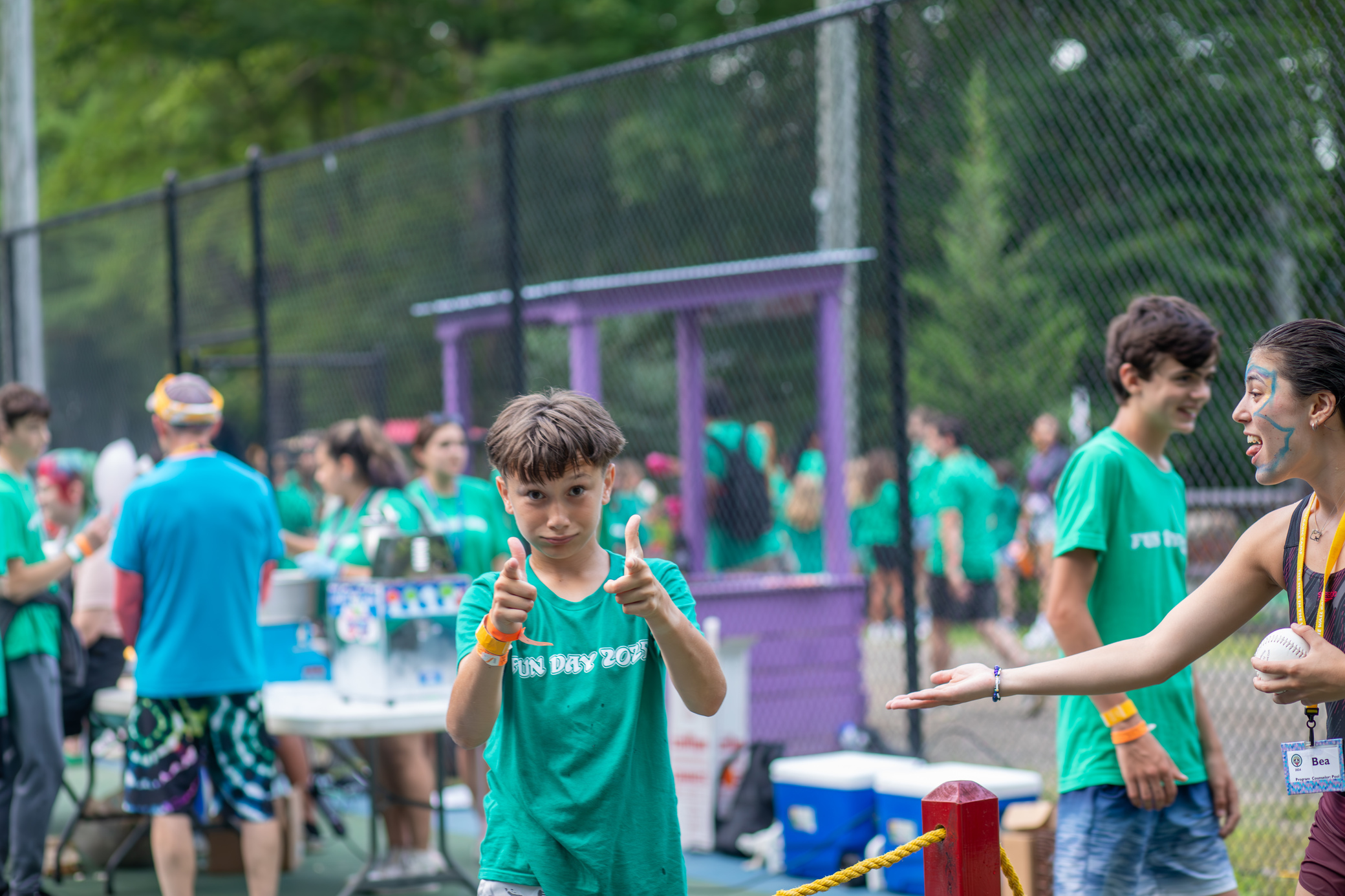 A boy in a FUN DAY 2023 shirt gives two thumbs up at an outdoor event. People in matching green shirts stand in the background near tables and a purple booth.
