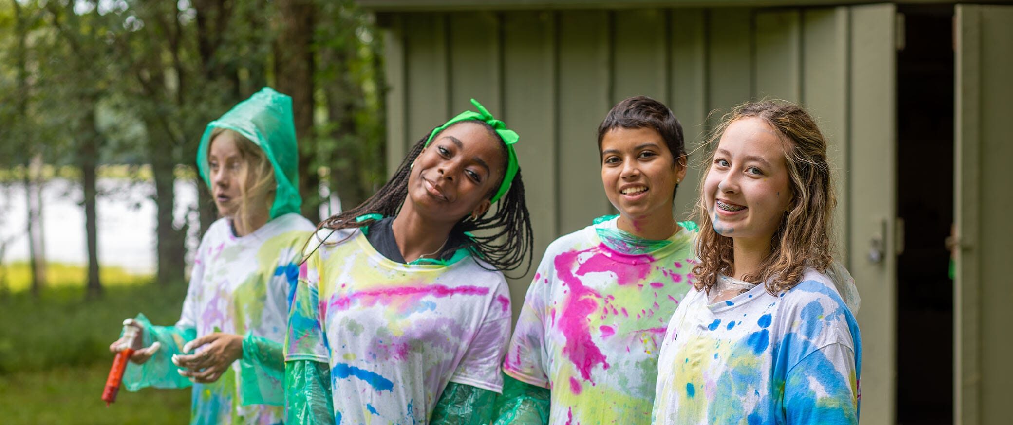 Four smiling kids in colorful, paint-splattered shirts stand outside near a green building. Trees and grass are in the background, suggesting a fun outdoor activity or camp event.