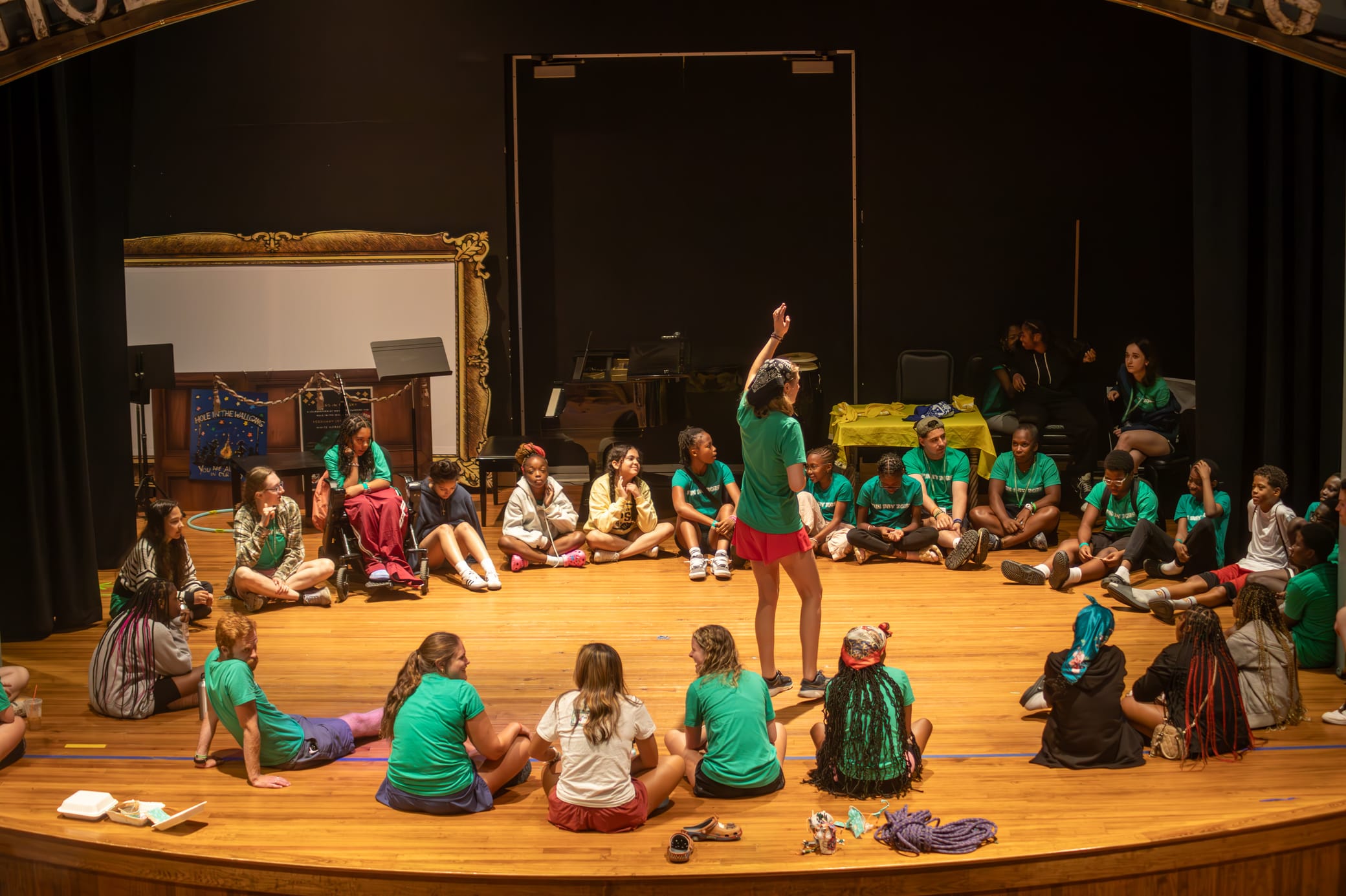 A group of young people in green shirts sit in a circle on a stage, listening to a person standing in the center with one arm raised. The setting is brightly lit with stage props in the background.