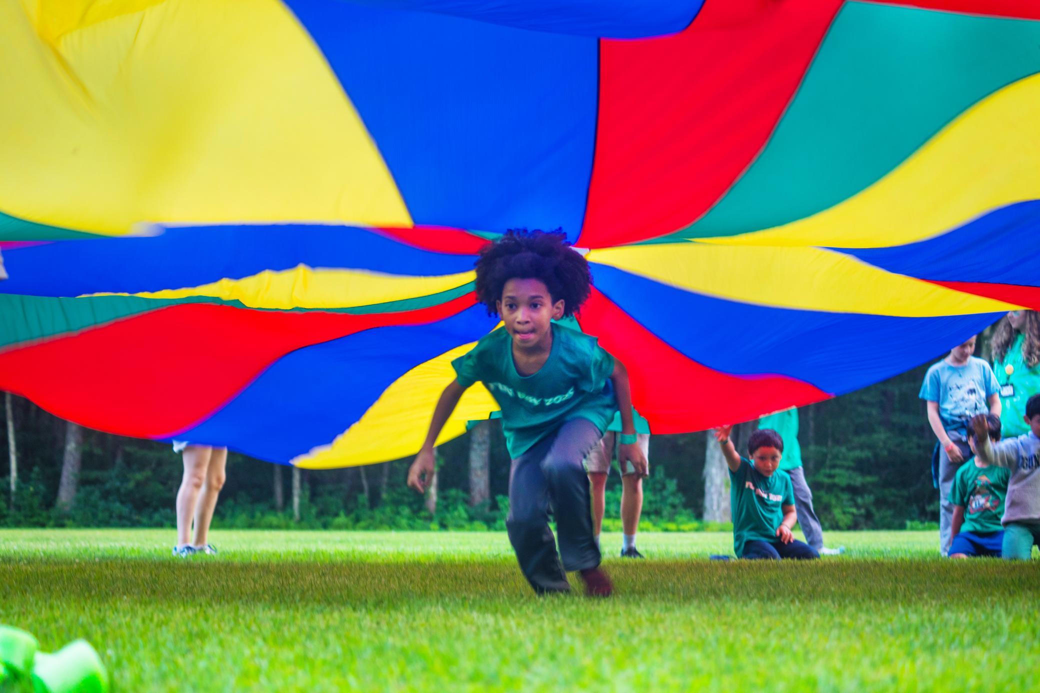 Children wearing green shirts play outside, crawling under a colorful parachute spread out above them on the grass, with trees in the background.