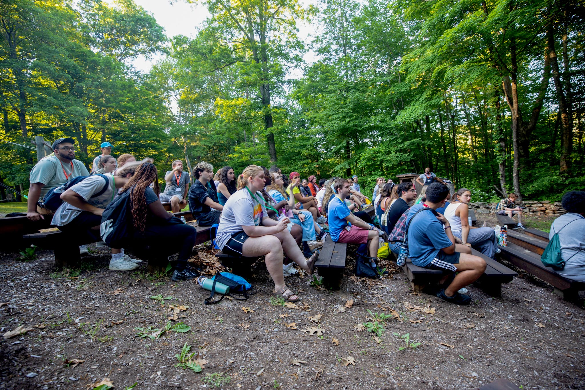 A group of people sit on wooden benches outdoors in a forest, attentively watching something out of view. Sunlight filters through green trees, and some people have backpacks by their feet.