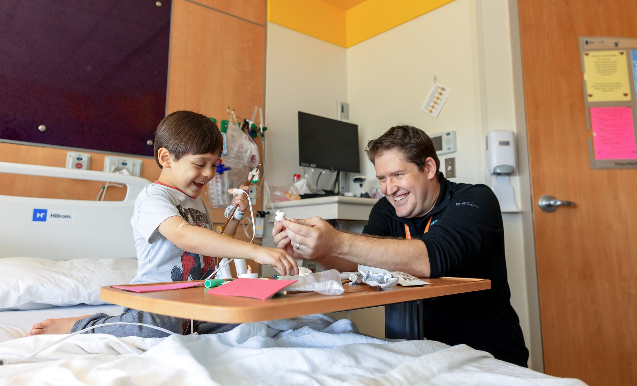A young boy in a hospital bed plays joyfully with a man at his bedside. Art supplies and paper are on a tray table, and medical equipment is visible in the room. Both appear happy and engaged.