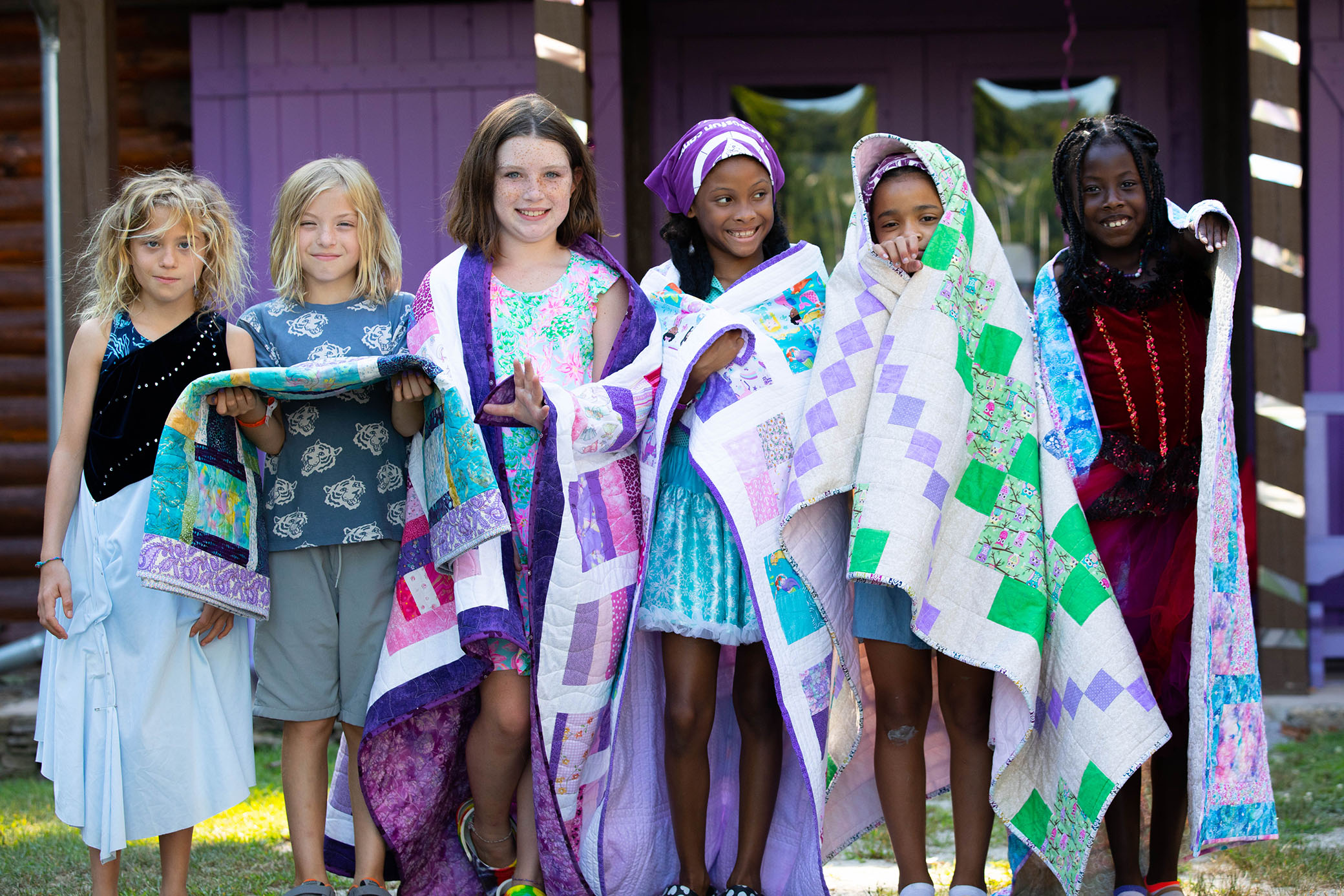 Six smiling children stand outside holding or wearing colorful quilts. They pose together in front of a purple painted wooden building, each showing off their unique quilt and outfits.