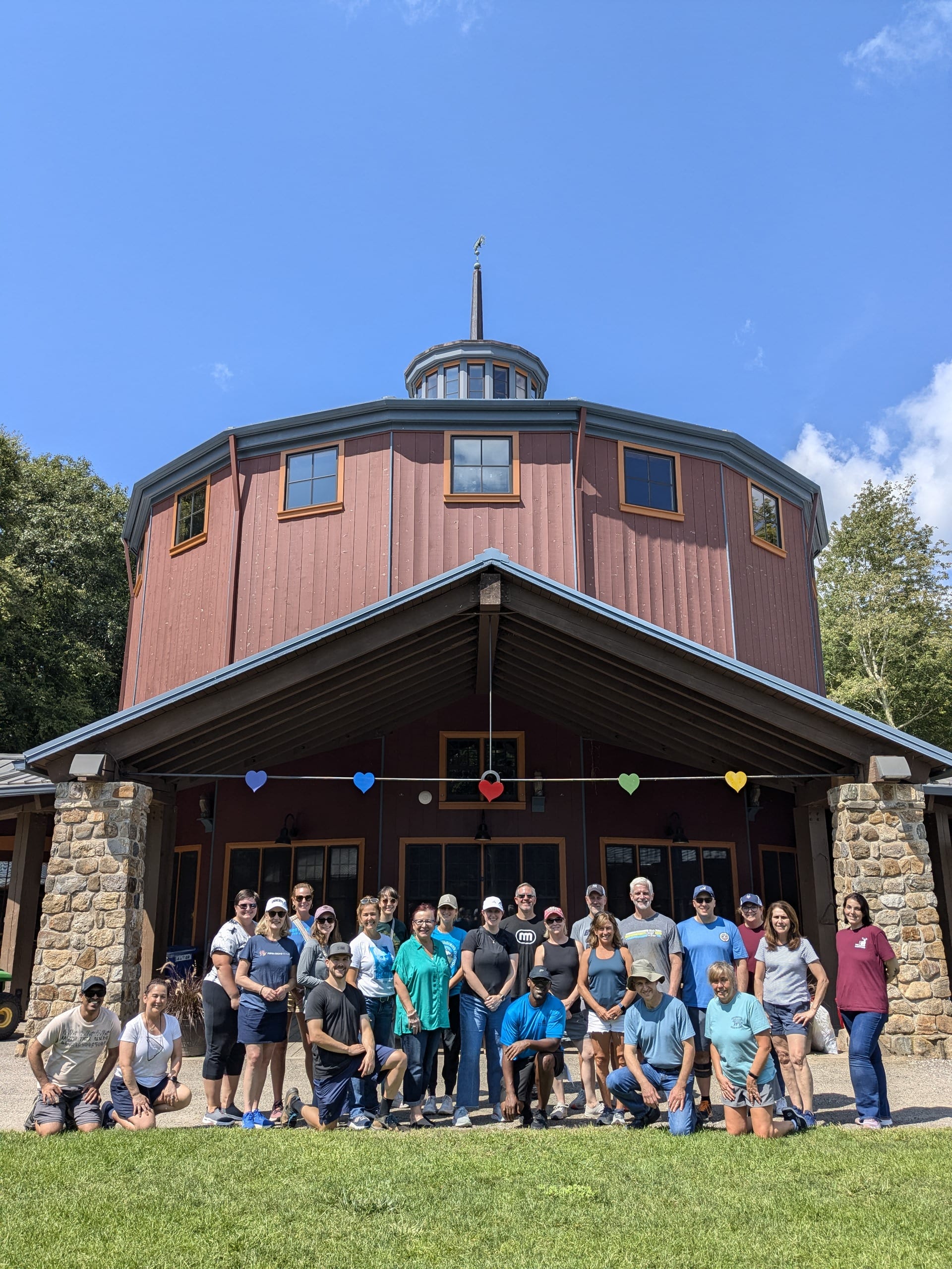 A group of people pose for a photo in front of a large, round, red building with stone pillars under a blue sky. Colorful heart decorations hang across the porch.