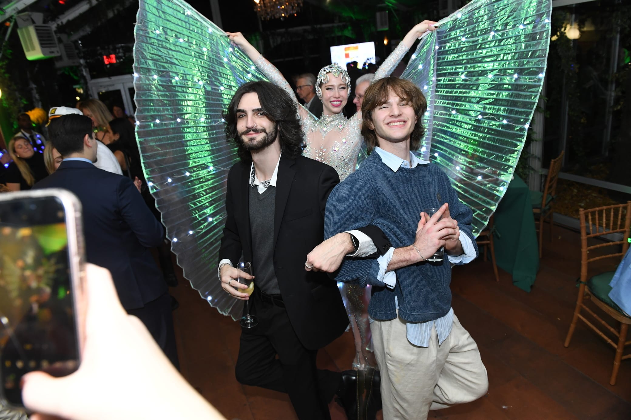 Two young men pose arm in arm at a party, smiling. Behind them stands a performer in a sparkling costume with large, illuminated wings, creating a festive atmosphere. Other guests are visible and someone is taking a photo.