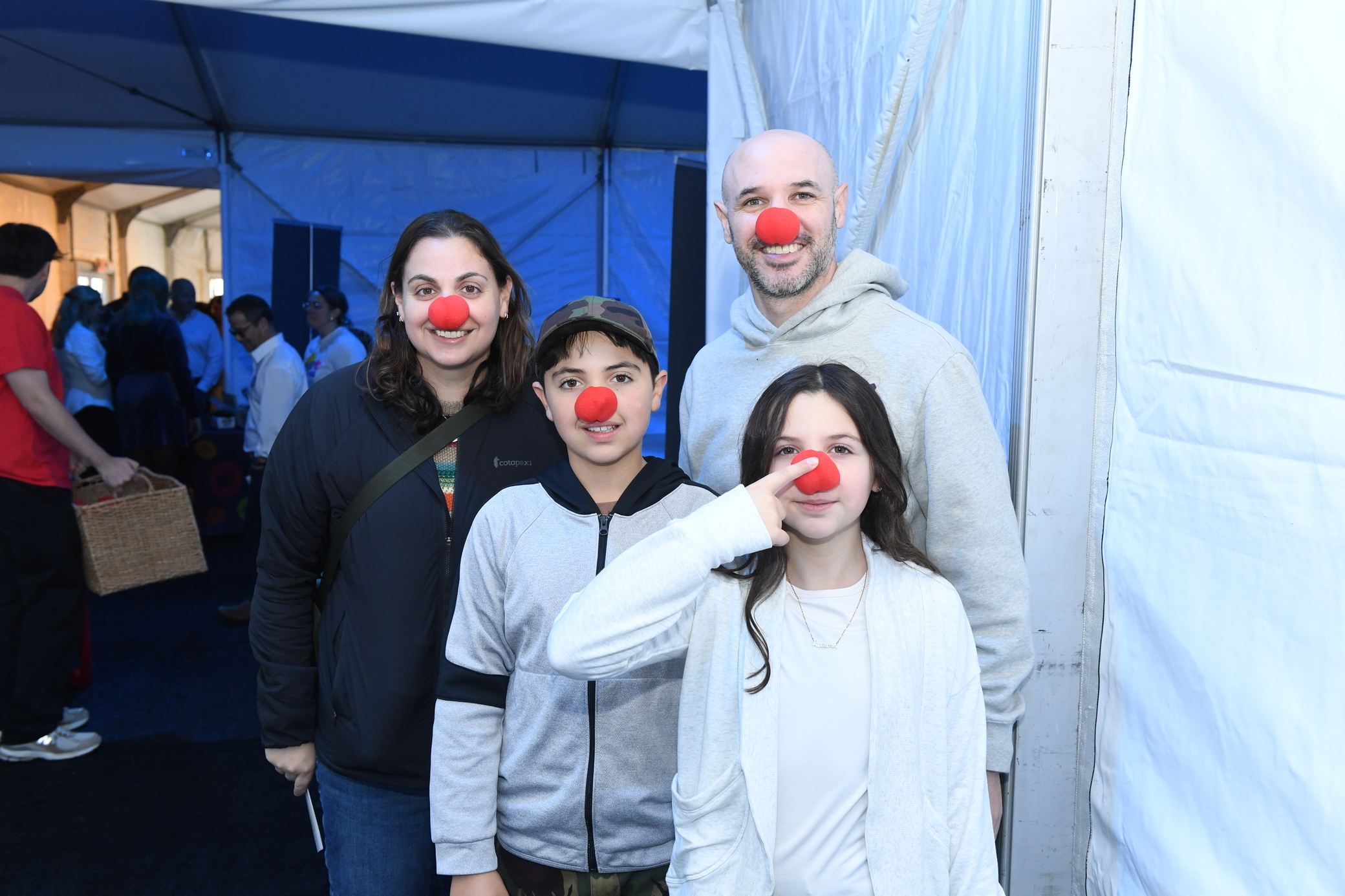 A smiling family of four, all wearing red clown noses, stands together inside a tent at an event.